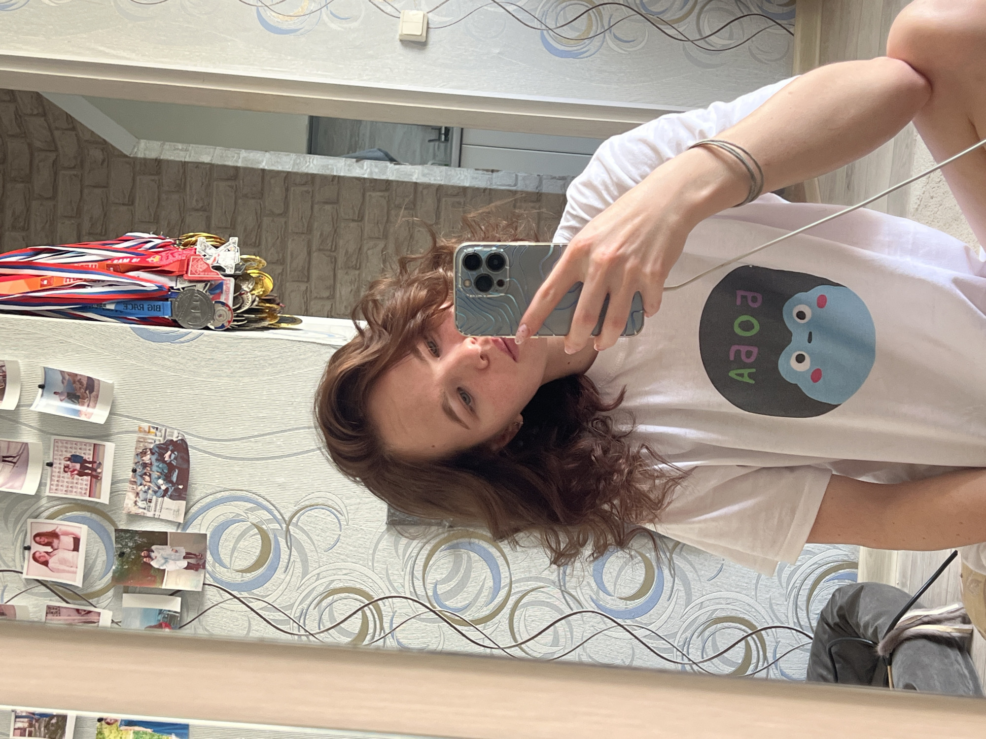A young woman takes a mirror selfie indoors, in what appears to be a bedroom or living area in Chaikovsky, Russia. The image is oriented horizontally, showing the woman with wavy brown hair, looking directly at the camera with a neutral expression. She is wearing a white t-shirt featuring a blue cartoon character and reversed Cyrillic text that appears to be "Я OAA" or "Я OPA" (likely "ААО Я" or "АРОЯ" when viewed directly). She holds an iPhone with a distinct blue, textured, wave-patterned case. A charging cable is draped over her arm.
The background features patterned wallpaper with swirling designs in blue, gold, and grey. Above the wallpaper, there's a plain light-colored wall with a white light switch. To the left of the frame, a collection of medals with red, white, and blue ribbons is stacked, indicating competitive achievements. One visible ribbon displays the reversed English text "ƎƆAЯ ⅁Iꓭ" (BIG RACE). Several small, framed photographs, depicting various people and group shots, are affixed to the wall. The overall lighting suggests it is daytime. The medal ribbons resembling the Russian flag colors and the Cyrillic text on her shirt are consistent with the provided location context of Russia.