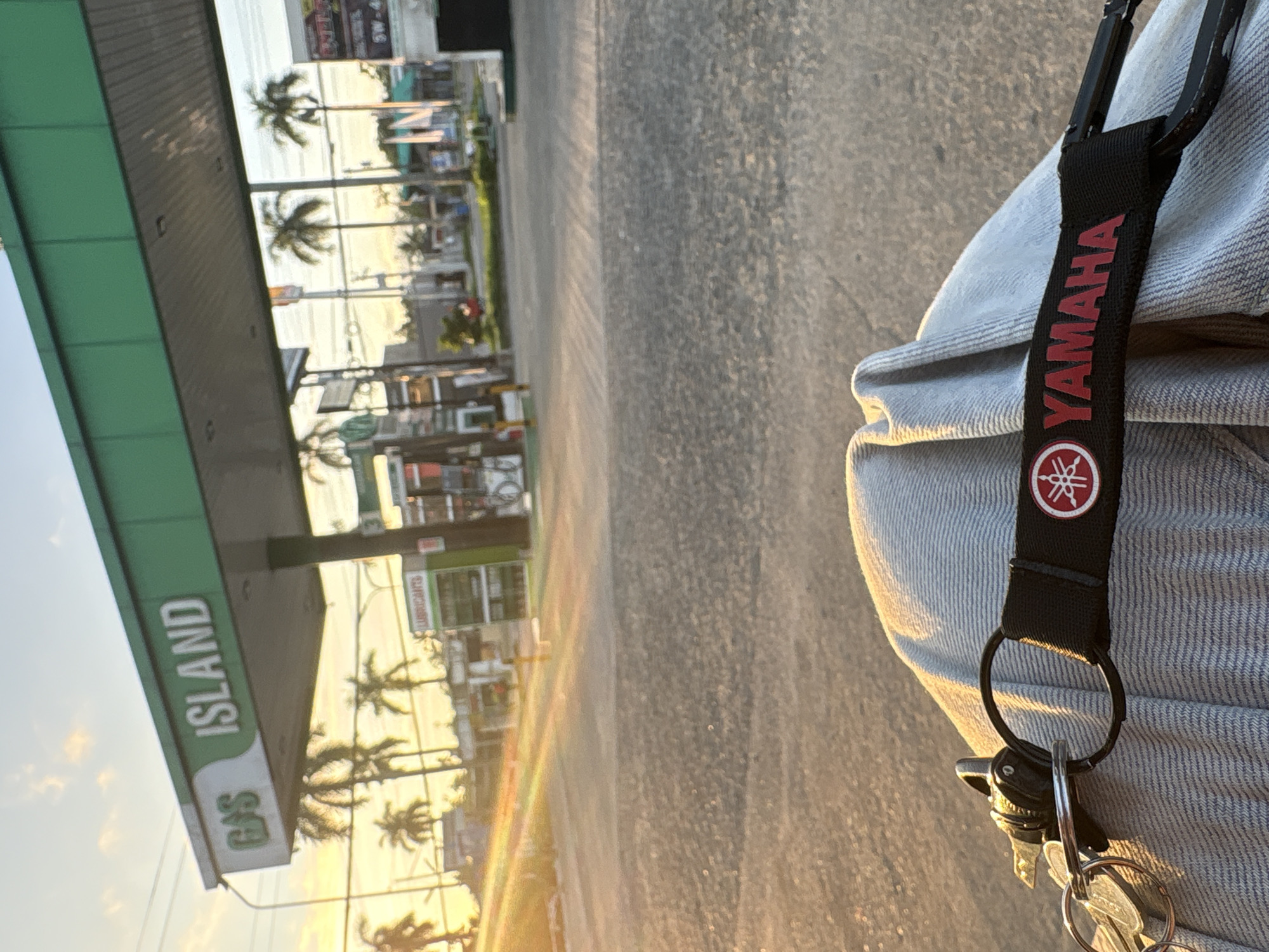 The image shows a gas station in Tagum City, Philippines, with the "GAS ISLAND" sign clearly visible. The sun is setting, casting a warm glow over the scene and creating lens flares. In the foreground, a Yamaha keychain is attached to a set of keys, resting on a person's lap, suggesting they are seated. Palm trees are scattered throughout the background, typical of the tropical climate in Tagum. The gas station appears to be operational, with pumps and a store visible. The overall atmosphere is serene, capturing a moment of pause at a local landmark.