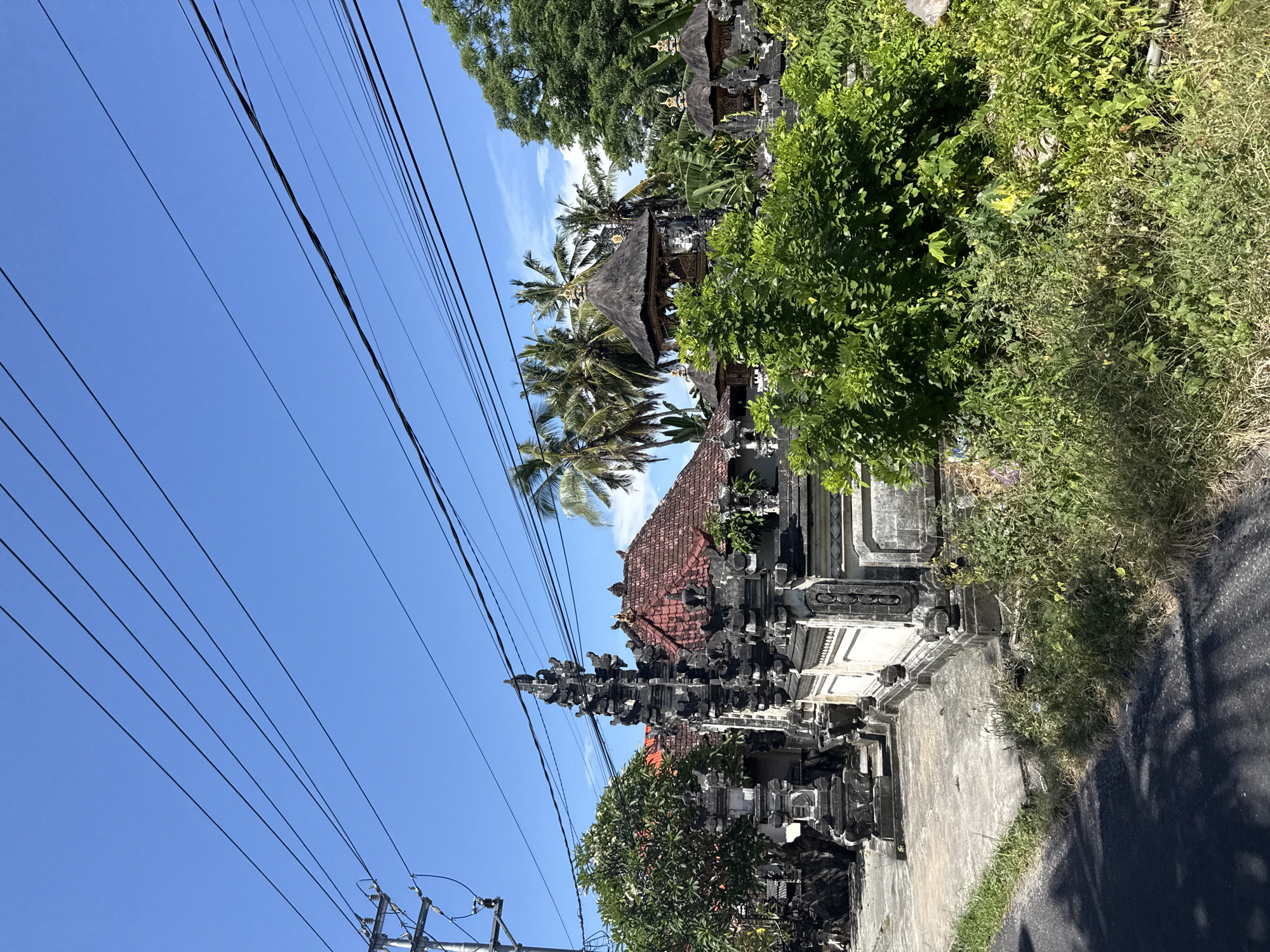 The image shows a street scene in Batununggal, Indonesia. A traditional Balinese gate structure, made of gray stone with intricate carvings, stands on the right side of the road. The gate is flanked by lush green bushes and other vegetation. Behind the gate, a building with a red-tiled roof and a thatched-roof structure can be seen among palm trees. In the background, the sky is a clear blue with a few wispy clouds. Power lines run across the sky in the upper left corner. The road is paved and appears to be lightly traveled.