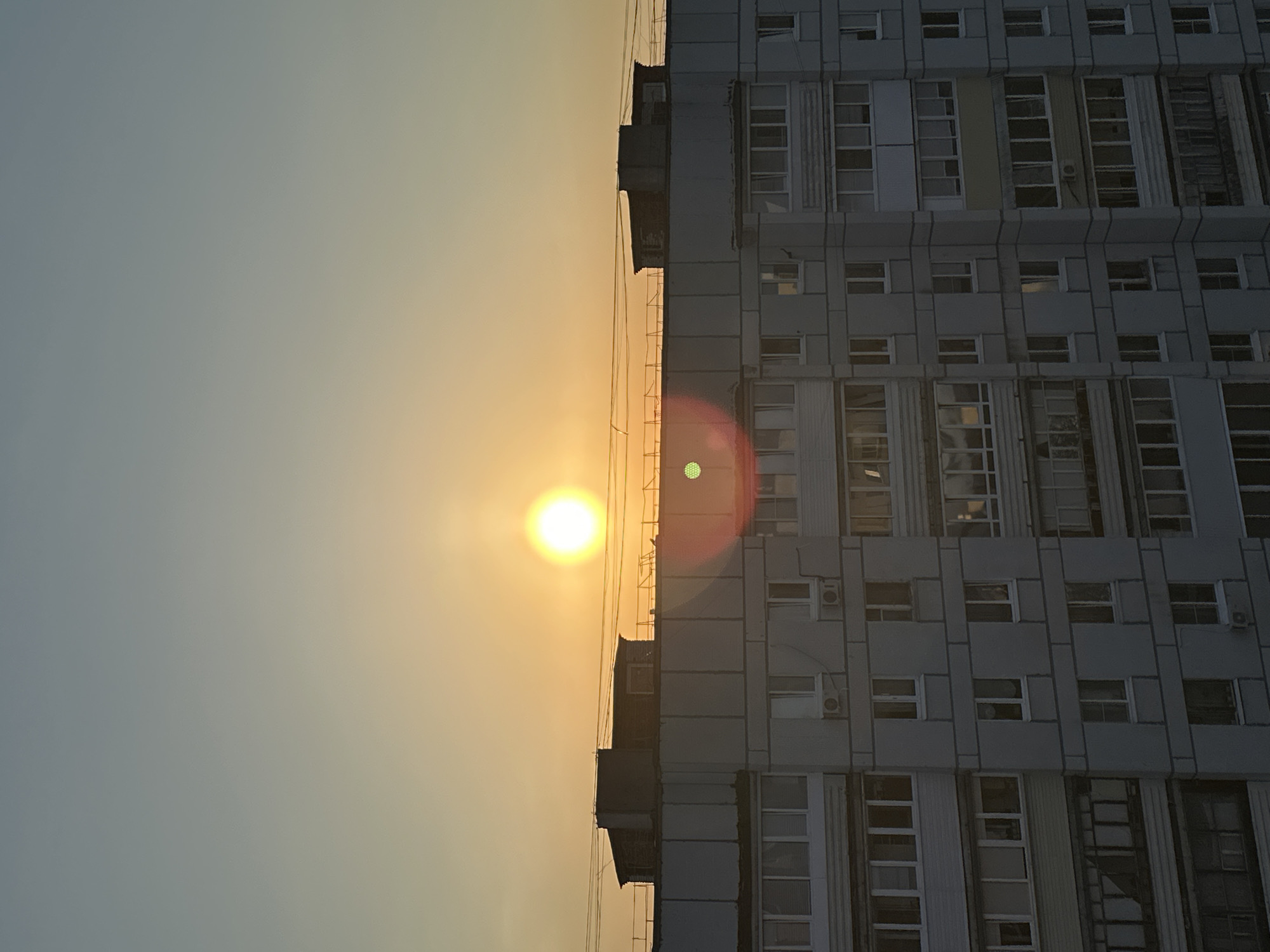 The image depicts a building facade, likely a residential or office structure, situated against a backdrop of a bright sunset. The building is rectangular and appears to be composed of modular units, each featuring multiple windows. The facade is a light gray color and seems to have a somewhat worn or aged appearance. In the foreground, the sun is positioned along the edge of the building, creating a strong contrast between the shadowed building and the brilliant sky. The image is oriented vertically, with the sun positioned towards the top-center and the building stretching downwards. Based on the provided data, this image is located in the city of Chaikovsky, Russia.