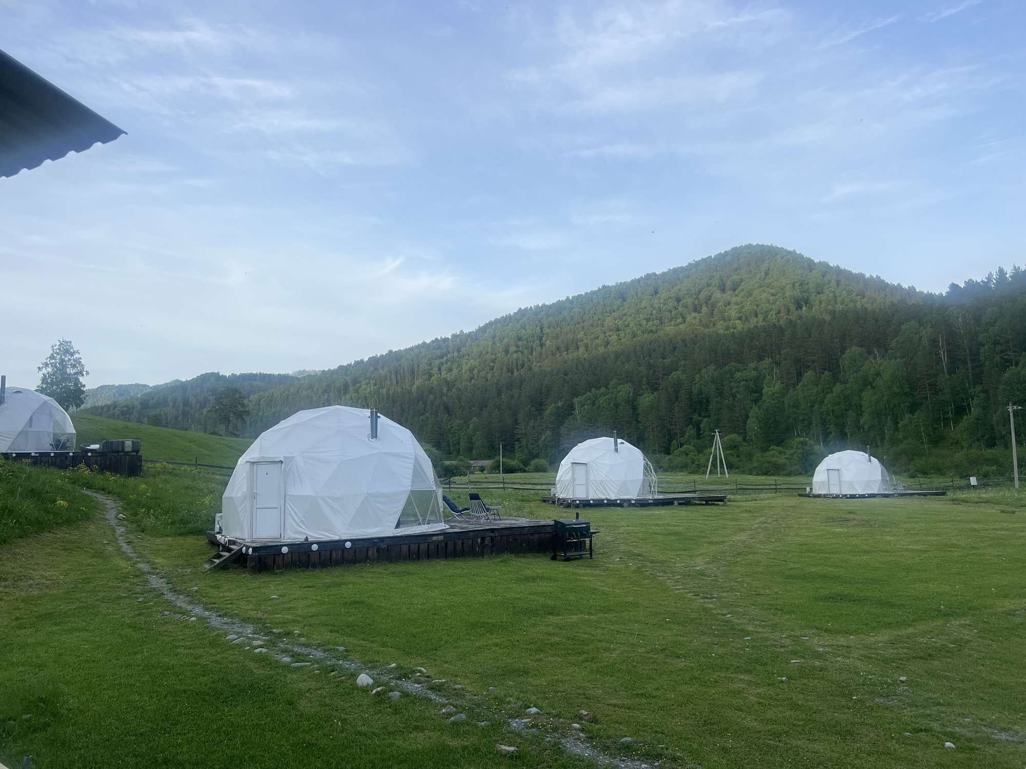 This image, captured in Uznezya, Russia, presents a glamping site featuring five white geodesic dome tents. The scene is dominated by a vast green grassy field under a pale blue sky with scattered thin white clouds.
In the foreground and mid-ground, five white geodesic dome tents are visible, each positioned on a dark-colored wooden platform. The closest dome, located on the left, has a white rectangular door at its front and a small set of wooden steps leading up to it. A dark metal pipe protrudes from its apex. Behind this dome and slightly to its left, another partial dome is visible. Towards the middle-right of the frame, a third dome tent stands on a similar platform, also featuring a white door and a metal pipe. A dark blue reclining chair is placed on this platform. In the grass directly in front of this dome, a dark-colored mobile barbecue grill is positioned. Further into the background on the right, two additional dome tents are discernible, each with a white door and a pipe, resting on platforms.
A narrow, winding path, composed of small stones and compacted earth, traverses the grassy field from the lower-center of the frame, leading towards the dome tents on the left. Small patches of yellow flora are interspersed within the green grass.
The background is characterized by a series of rolling hills densely covered with dark green coniferous and deciduous trees. In the distant background, amidst the tree line, utility poles with power lines are faintly visible. The roofline of a dark-colored structure is partially visible in the upper-left corner of the image. No individuals are present in the scene. The natural light suggests a daytime setting, possibly morning or late afternoon, with soft, ambient illumination.