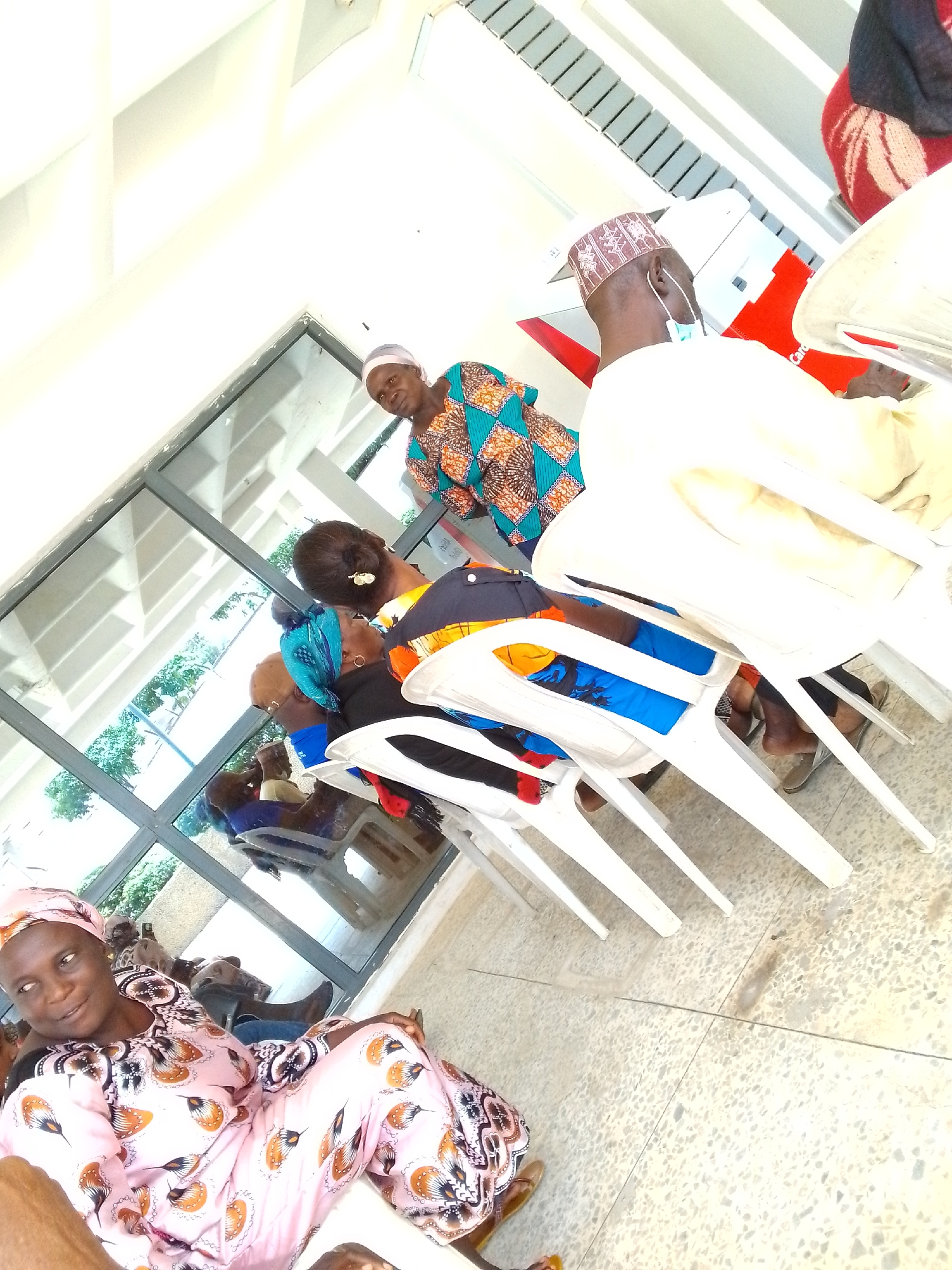 In Jos, Nigeria, a group of people are gathered indoors, possibly in a waiting area or community space. The scene is brightly lit, with natural light filtering in through large windows.
In the foreground, a woman wearing a light pink and patterned traditional outfit is seated, looking towards the left side of the frame with a slight smile. Her head is covered with a matching headscarf.
Behind her, several other individuals are visible. A woman in the mid-ground is wearing a vibrant blue, green, and orange patterned garment and a light-colored headscarf. She is looking down and slightly to the left. To her right, a man is seated, facing away from the camera. He is wearing a light-colored traditional cap with a pattern and a light-colored garment. A light-colored mask is worn below his chin. He appears to be wearing earphones.
Further back, other people are seated or standing. The environment features a speckled, light-colored floor. White plastic chairs are arranged, with some occupied by people and others empty. Reflections are visible in the large windows, showing some outdoor greenery. In the background, a red and white structure, possibly part of an advertisement or a building feature, is partially visible. The overall impression is one of a casual gathering.