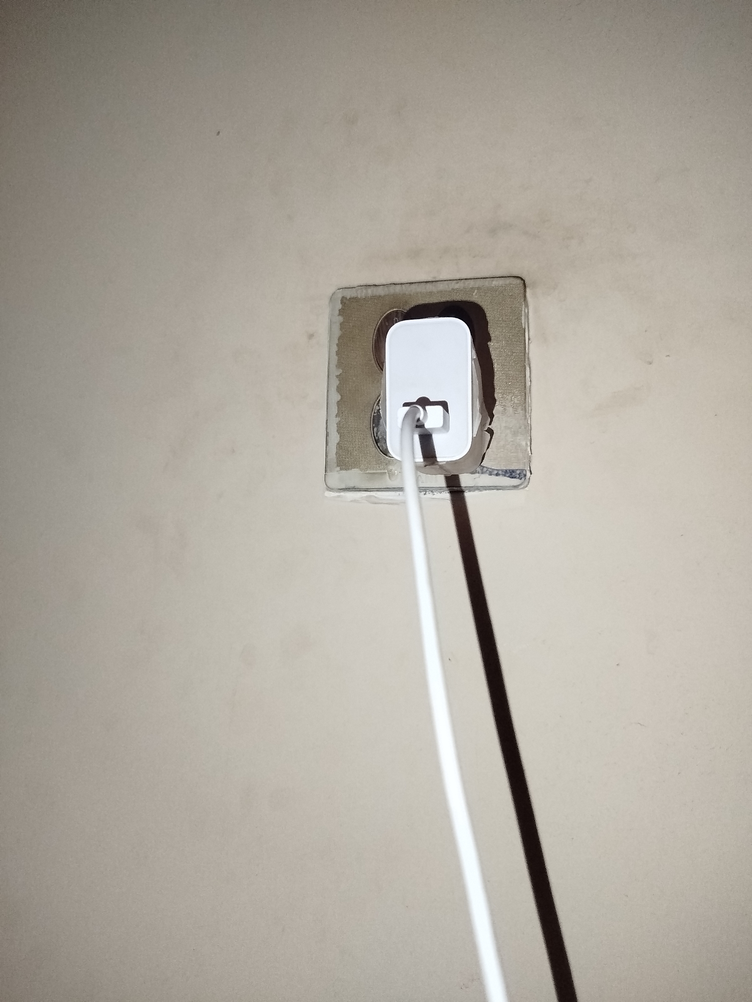 A white power adapter is plugged into a wall socket, with a white cable extending from it. The wall surrounding the socket appears to be textured and lightly discolored, with some faint marks and patches visible. The lighting suggests an indoor environment, casting a shadow from the cable. The specific location in Lassan, Pakistan, cannot be determined from the visual information provided.