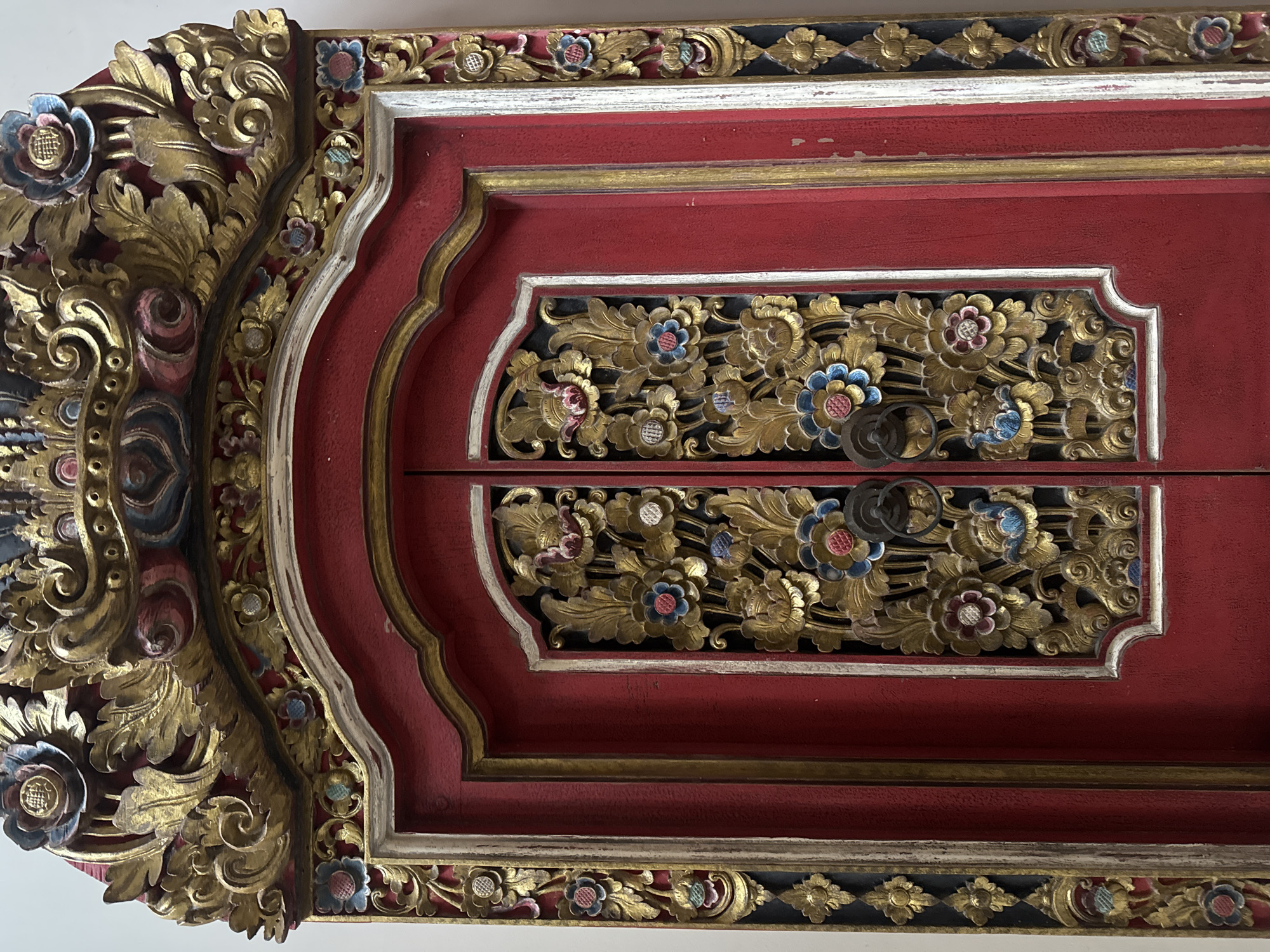 This is a close-up, eye-level, indoor photograph of a red, ornately carved wooden door with gold accents. The carving features intricate floral and foliage patterns, with some elements painted in blue, pink, and white. The door has two panels, each with a circular metal handle. The door is framed by a decorative border, also featuring carvings and gold accents, with vertical segments on either side of the door. The background is a plain white wall. This door appears to be located in Denpasar, Indonesia, potentially as an architectural feature of a traditional Balinese building or a piece of decorative furniture.