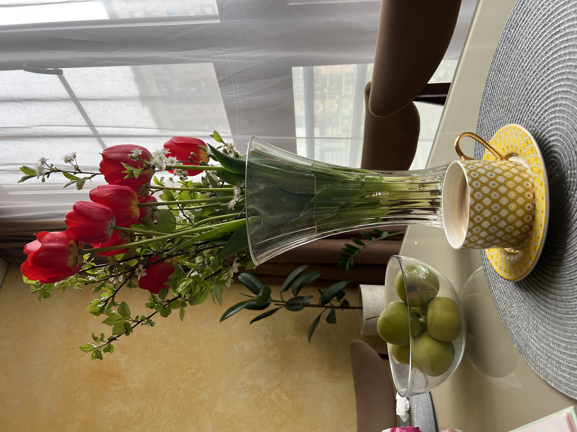 The image shows a vase of red tulips with green leaves and white flowers on a table. Next to the vase is a glass bowl filled with green apples. A yellow and white patterned teacup sits on a matching saucer, next to a grey placemat. The background features a window with white blinds, and a chair. The location is not discernible from the image. It is not possible to confirm a location in Khimki, Russia.