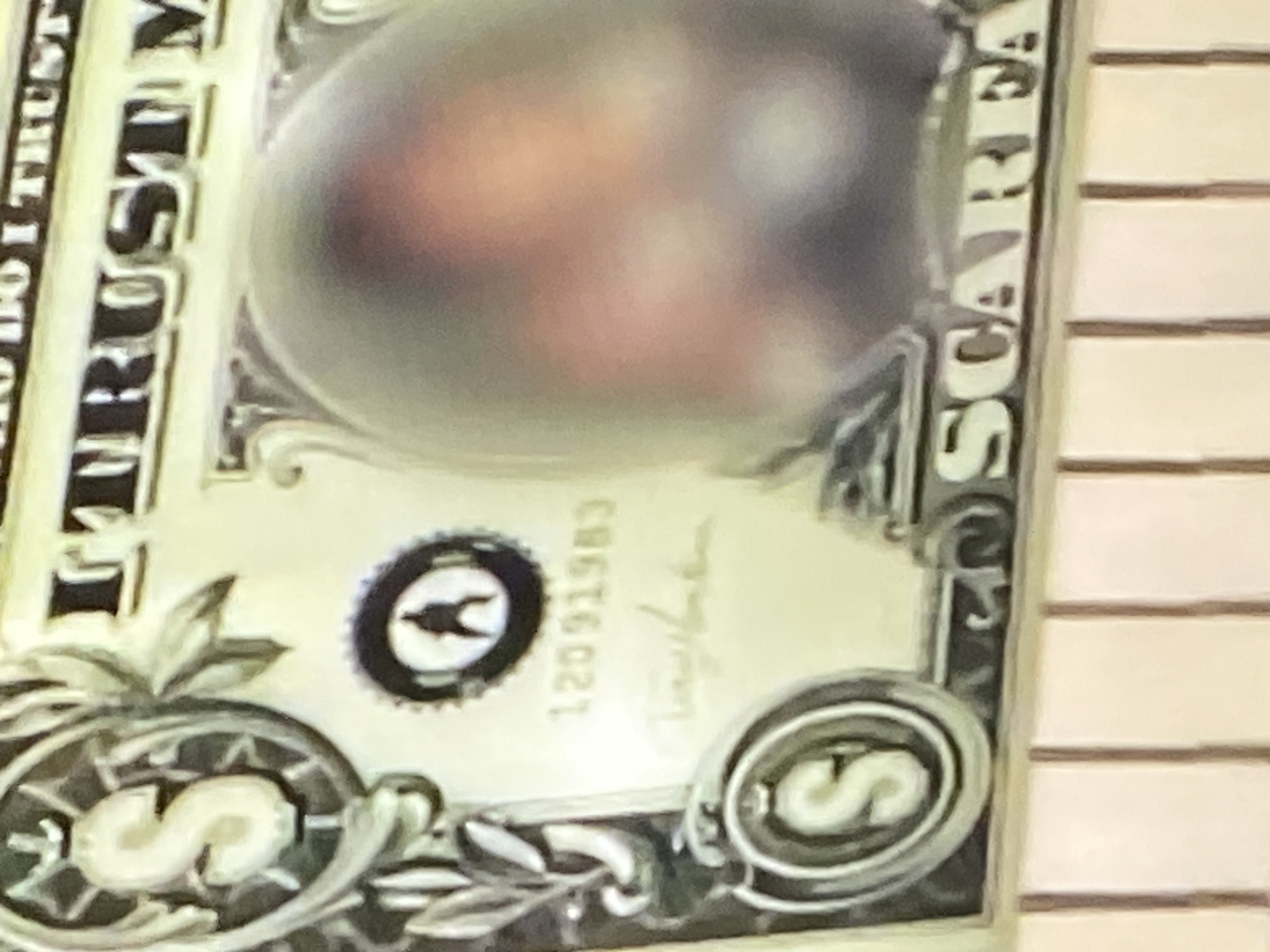 The image shows a close-up of a one-dollar bill, partially obscured by what appears to be a reflective, bubble-like object. The bill is positioned horizontally, with the top edge showing the words "IN GOD WE TRUST" and "THE UNITED STATES OF AMERICA". To the left, a large letter "S" is visible within a circular design. Below the "S", there's a seal with a silhouette of a person. Further to the right, a serial number "12091983" is faintly visible, followed by a handwritten signature. The most prominent feature is the large, blurry, oval-shaped object in the center, which has a reddish-brown hue and suggests the presence of a face or faces within it. On the bottom right of the bill, the word "SCARFACE" is clearly printed in large letters. The background appears to be a textured, light-colored surface, possibly a table or shelf, with vertical lines indicating some sort of structure or separation.
This image cannot be geolocated to Yekaterinburg, Russia, as it depicts a U.S. dollar bill and contains no identifiable elements related to that city.