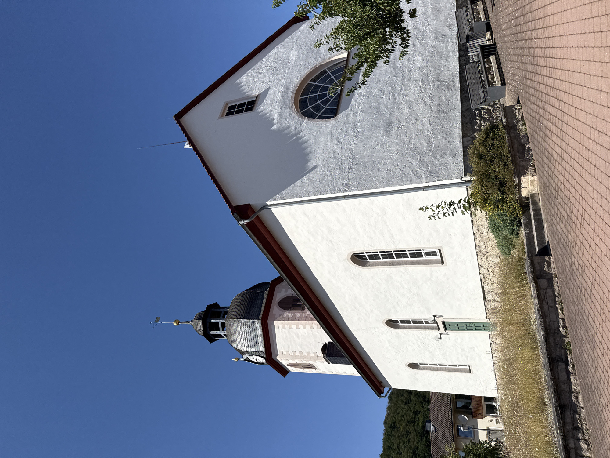 A white church with a dark gray, multi-tiered roof and a clock tower with a dark gray, conical roof topped with a golden weather vane stands under a clear blue sky. The church has several tall, arched windows with white frames and one large, arched window with a grid pattern on the right side. A green door is visible on the left side of the building. The church is situated in a grassy area with a stone foundation. A tree with green leaves partially obscures the right side of the church. In the foreground, a red brick pathway leads up to the church. In the background, a wooded hill and a residential building are visible. This is the evangelische Kirche St. Salvator in Bad Sachsa, Germany.