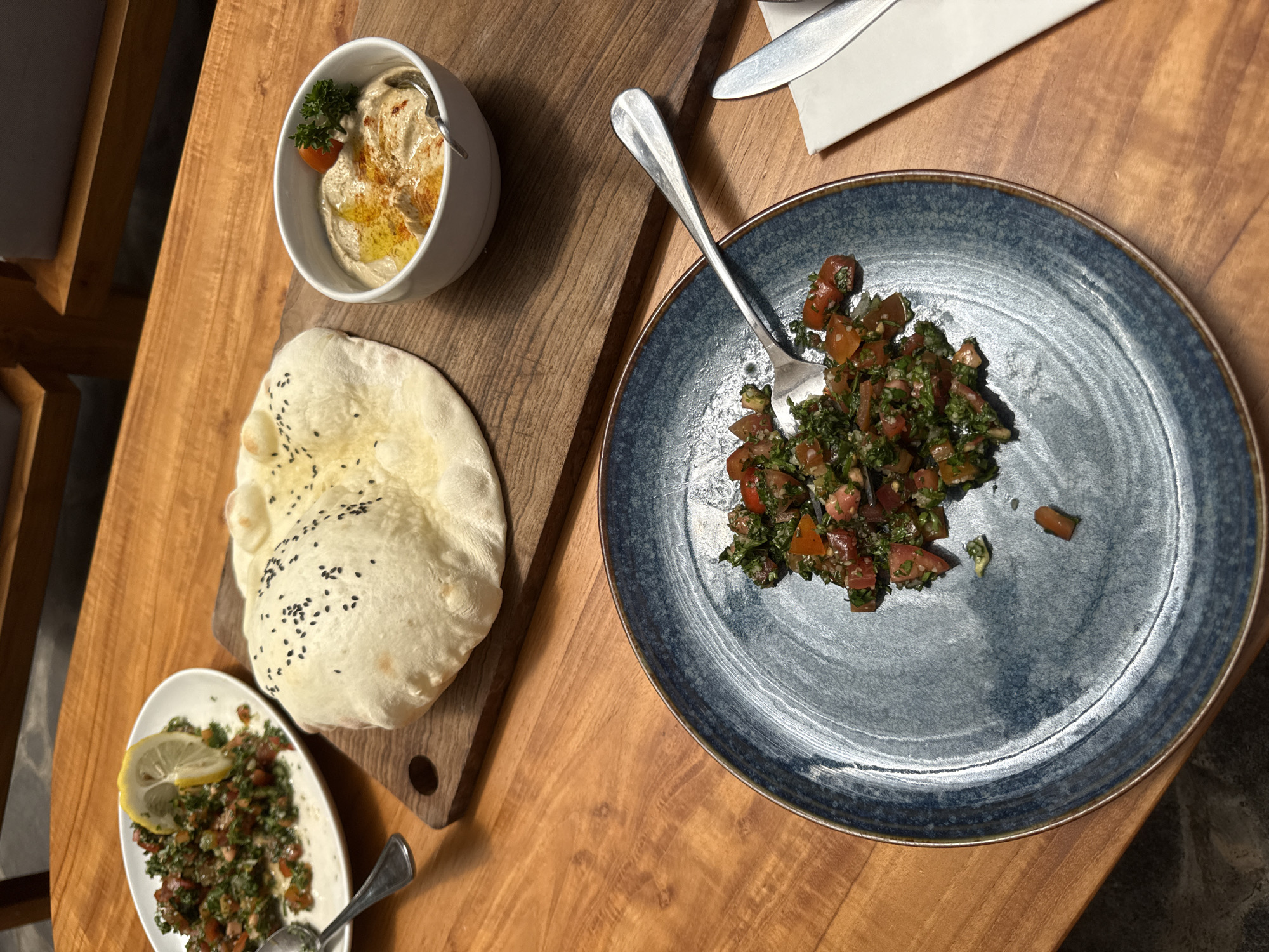 This image presents an overhead view of a wooden table laden with a selection of Middle Eastern or Mediterranean dishes, likely served in a restaurant setting in Ubud District, Indonesia.
The scene is dominated by various food items meticulously arranged. In the upper left, a white bowl holds a generous portion of hummus, artfully garnished with a sprig of green herb, a small red cherry tomato, a drizzle of olive oil, and a sprinkle of reddish spice, possibly paprika or sumac. A spoon rests within the hummus, ready for serving. Adjacent to this, on a rustic wooden cutting board, lies a large, round piece of freshly baked flatbread, characteristic of pita bread, generously adorned with black sesame seeds.
To the right, a textured blue ceramic plate showcases a portion of a finely chopped salad, appearing to be tabbouleh or a similar blend of herbs, tomatoes, and perhaps bulgur. A silver fork is positioned in the salad. Further down on the left side of the table, a smaller, white oval plate holds another serving of a similar herb and tomato salad, garnished with a bright yellow lemon slice. A spoon is also present in this dish.
In the upper right corner, a glimpse of a neatly folded white napkin encasing a knife and fork indicates a complete place setting. The wooden table exhibits a warm, natural grain, contributing to an inviting and earthy ambiance. A dark wooden chair or architectural detail is partially visible on the far left, complementing the overall decor. The soft, ambient lighting suggests an indoor setting, possibly an evening meal. No visible text is present in the media file.