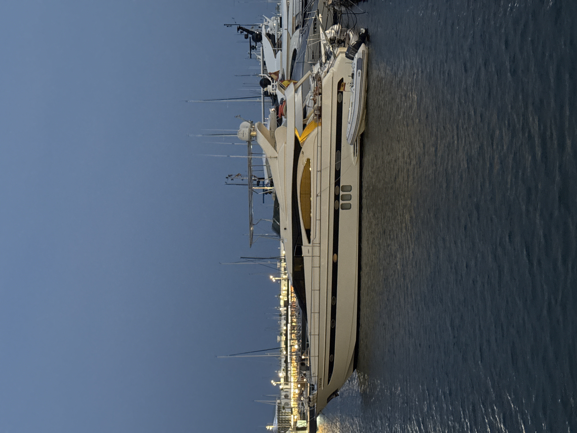 The image depicts a large, cream-colored yacht docked in a marina, likely at dusk or dawn, given the low light conditions and the illuminated buildings in the background. The yacht is the primary subject, positioned centrally and occupying a significant portion of the frame. It has a sleek design with dark trim along its hull and several oval windows. A smaller inflatable boat is tethered to the side of the yacht.
In the background, there are numerous other boats, including sailboats with masts visible. The marina area is lined with buildings that are illuminated, suggesting an urban or developed waterfront. The water in the foreground is dark and choppy, reflecting the ambient light.
The overall scene conveys a sense of quiet luxury and maritime activity. The sky is a deep blue-gray, indicating overcast or twilight conditions. The location is identified as the marina in Alicante, Spain. There are no people visible in the image.