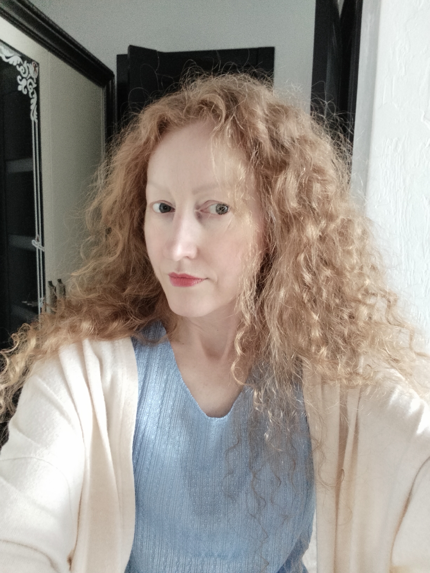 A woman with long, curly, strawberry-blonde hair is posing indoors. She has fair skin, light eyebrows, and appears to be wearing red lipstick. She is wearing a light blue, textured top and a cream-colored cardigan.
Behind her are two doorways or dark-colored framed openings. One doorway has a white decorative design. The wall to her right is white with visible texture. The exact location in Krykhivtsi, Ukraine, is not discernible from the image.