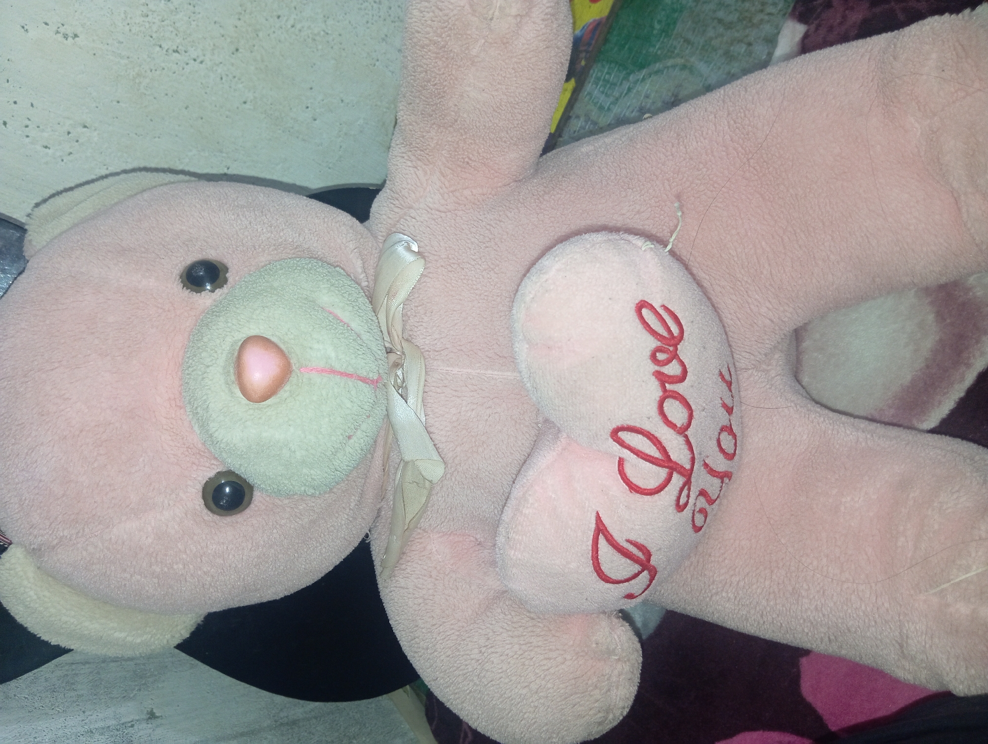 A close-up shot captures a plush pink teddy bear sitting upright. The teddy bear has a pale pink nose and a lighter pink muzzle. A beige ribbon is tied in a bow around its neck. In its hands, the teddy bear holds a pink heart-shaped pillow that is embroidered with the words "I Love You" in red cursive. The teddy bear is positioned in a seated posture, with its arms resting on its lap. The background is out of focus, but suggests an indoor setting, possibly a home environment in Rejosari, Indonesia. The lighting is dim, casting a soft glow on the teddy bear and its accessory.
