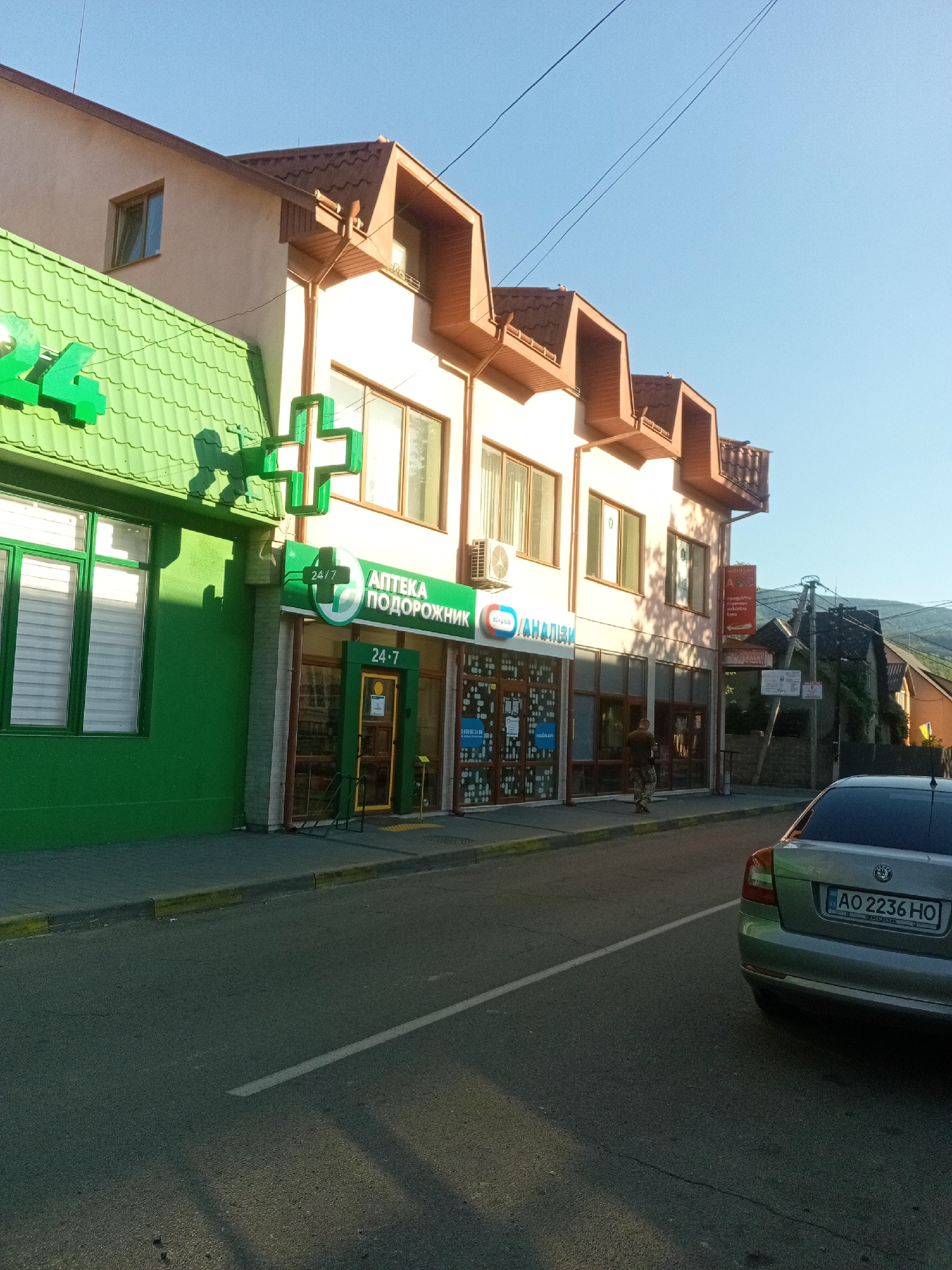 The image shows a street scene in Tiachiv, Ukraine. A green-fronted building with a large green cross sign and "24/7" lettering is on the left. Next to it is a beige building with multiple windows and dormer roofs, featuring signage that includes "24/7 АПТЕКА ПОДОРОЖНИК" and other text in Cyrillic. A person in camouflage clothing stands near the entrance of the beige building. A silver Skoda sedan with license plate AO 2236 HO is parked on the right side of the street, facing away from the camera. The street is paved with asphalt and has a white line marking. The sky is clear and blue. Overhead power lines are visible. The environment appears to be a commercial area in a town or city.