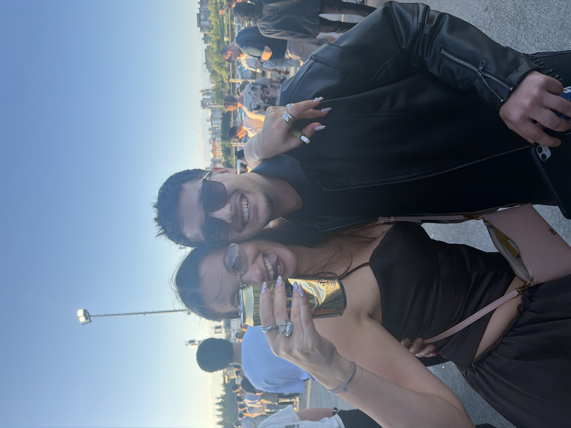The image shows a man and a woman smiling and posing closely together. The man wears sunglasses and a black jacket. The woman is holding a beverage can and wears sunglasses, a dark top, and a light pink purse. They are positioned in front of a crowd of people. Streetlights are visible in the background, and buildings are visible on the horizon. Based on the context provided, the location is in the city of Başakşehir, Turkey.