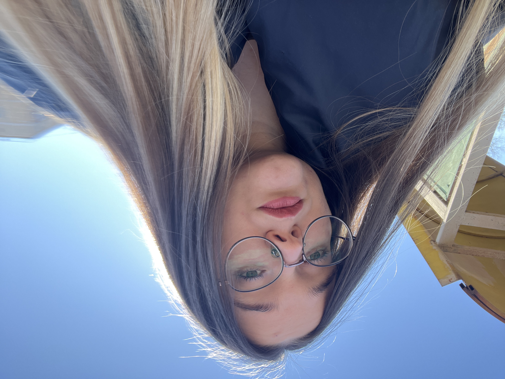 The image is a low-angle, head-and-shoulders selfie of a female. She possesses long, straight hair with prominent blonde highlights, strongly backlit on the right, creating a bright halo effect. Her facial features include light-colored eyes behind round, thin-framed eyeglasses, and lips colored with a reddish-pink tint. She is wearing dark-colored attire, possibly a jacket. Her gaze is directed straight at the camera with a neutral expression. The background features a clear, bright blue sky dominating the upper right. In the upper left, a segment of a yellow-painted structure, possibly an overhang or eave with visible support beams, is present, alongside a faint glimpse of a light green wall section below it. The lighting indicates an outdoor daytime setting under direct sunlight. The specific location within Bol'shevik, Russia, is not identifiable from the visual information contained within the image.