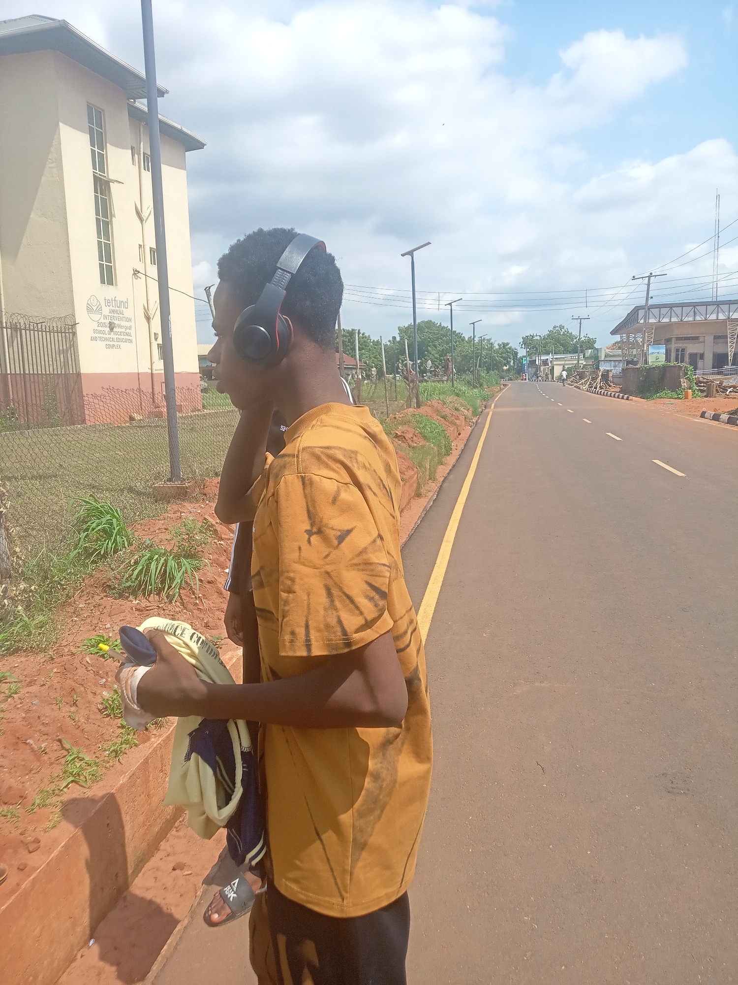An outdoor, eye-level shot captures a young person standing by the side of a paved road in Nsugbe, Nigeria, likely during the daytime. The subject, facing left with a slight profile, wears a yellow/orange tie-dye t-shirt, dark shorts, and black over-ear headphones with red accents. Black slides with "PEAK" visible on them are on their feet, and they hold a light-colored piece of clothing or fabric in their right hand. Another person is partially visible behind the main subject.
To the left, behind the subjects, stands a light-colored institutional building with tall, narrow windows and a prominent sign. The sign reads: "tetfund", "ANNUAL INTERVENTION", "2015/ Merged", and "SCHOOL OF VOCATIONAL AND TECHNICAL EDUCATION COMPLEX". A metal fence runs in front of the building, separated from the roadside by reddish-brown dirt and sparse green plants.
The road features a solid yellow line marking the edge of the shoulder, and further into the asphalt lane, broken white lines are visible. Along the road, several street lights or utility poles are visible. In the distance on the right, another building, possibly under construction, can be seen. The sky above is partly cloudy, suggesting bright, fair weather. The overall setting depicts a street adjacent to an educational or vocational complex.