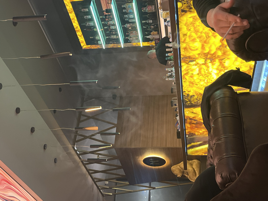 The image displays the interior of a commercial establishment, likely a lounge or bar, located in Burgas, Bulgaria. In the middle ground, a bald male individual, wearing a dark shirt, stands with his back to the camera behind a bar counter. The bar's front is illuminated with a bright, warm yellow-orange light, showcasing a translucent, crystalline pattern. Behind the bartender, a back bar contains multiple shelves filled with various liquor bottles, illuminated by prominent horizontal teal light strips.
The foreground is occupied by dark brown, tufted leather armchairs. On the right, a person's hand is visible, holding a white cigarette or similar slender object, resting on the armrest of an armchair. A dark jacket or piece of clothing is draped over an adjacent armchair to the left.
The ceiling features multiple cylindrical pendant lights, some vertically oriented, providing localized illumination. Wisps of smoke or vapor are discernible in the air, particularly in the upper mid-ground. To the left of the main bar, a wooden-paneled structure with a geometric lattice design above it includes a backlit circular sign displaying the initials "NP." Below this sign, a gold-colored statue of a seated gorilla or ape is positioned on a ledge.