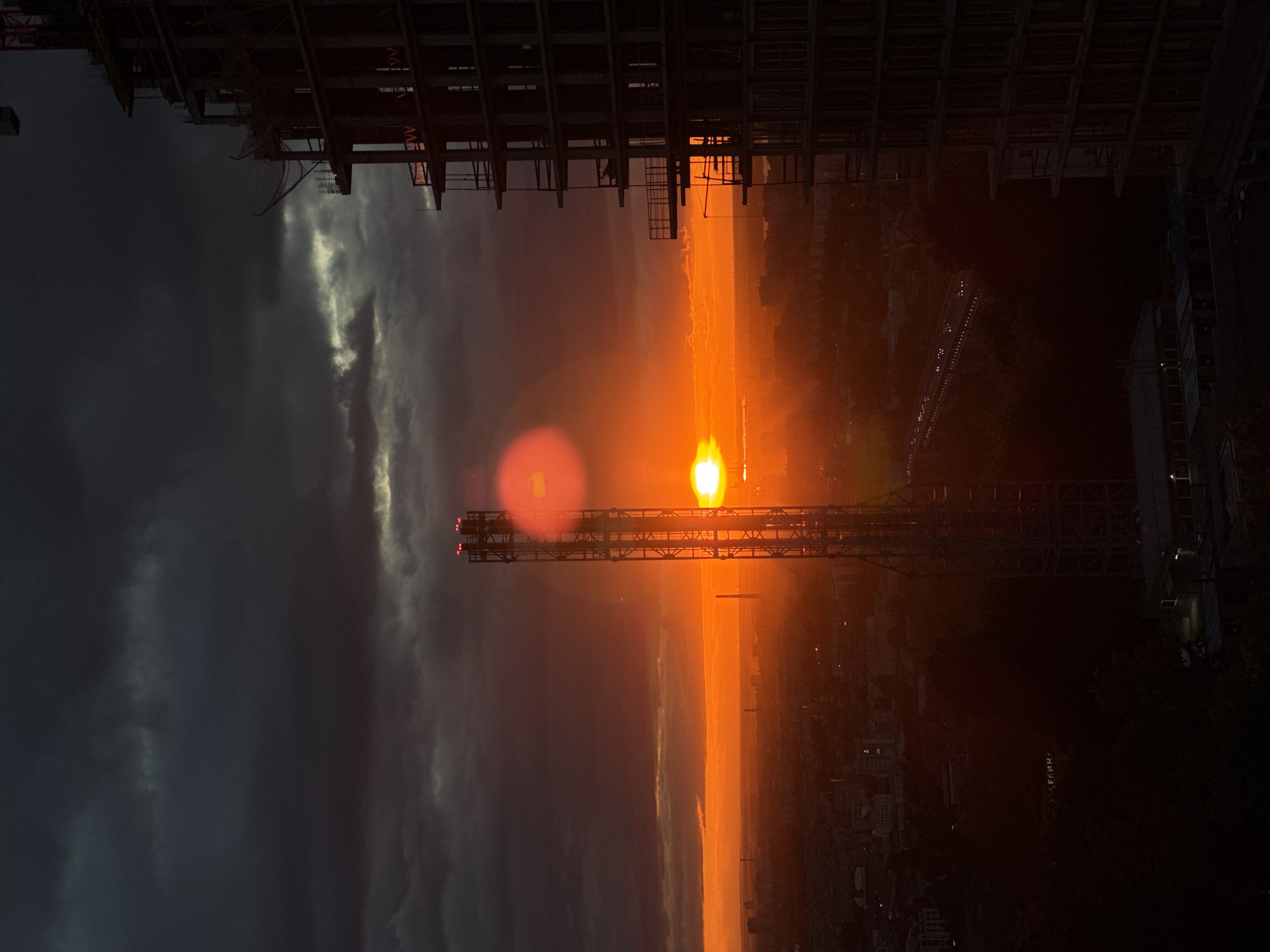 A construction site at sunset in an unknown city. A tall construction crane with lights runs horizontally across the center. Above and below are the dark frames of unfinished buildings. The sunset is bright orange. The left side of the image has storm clouds.