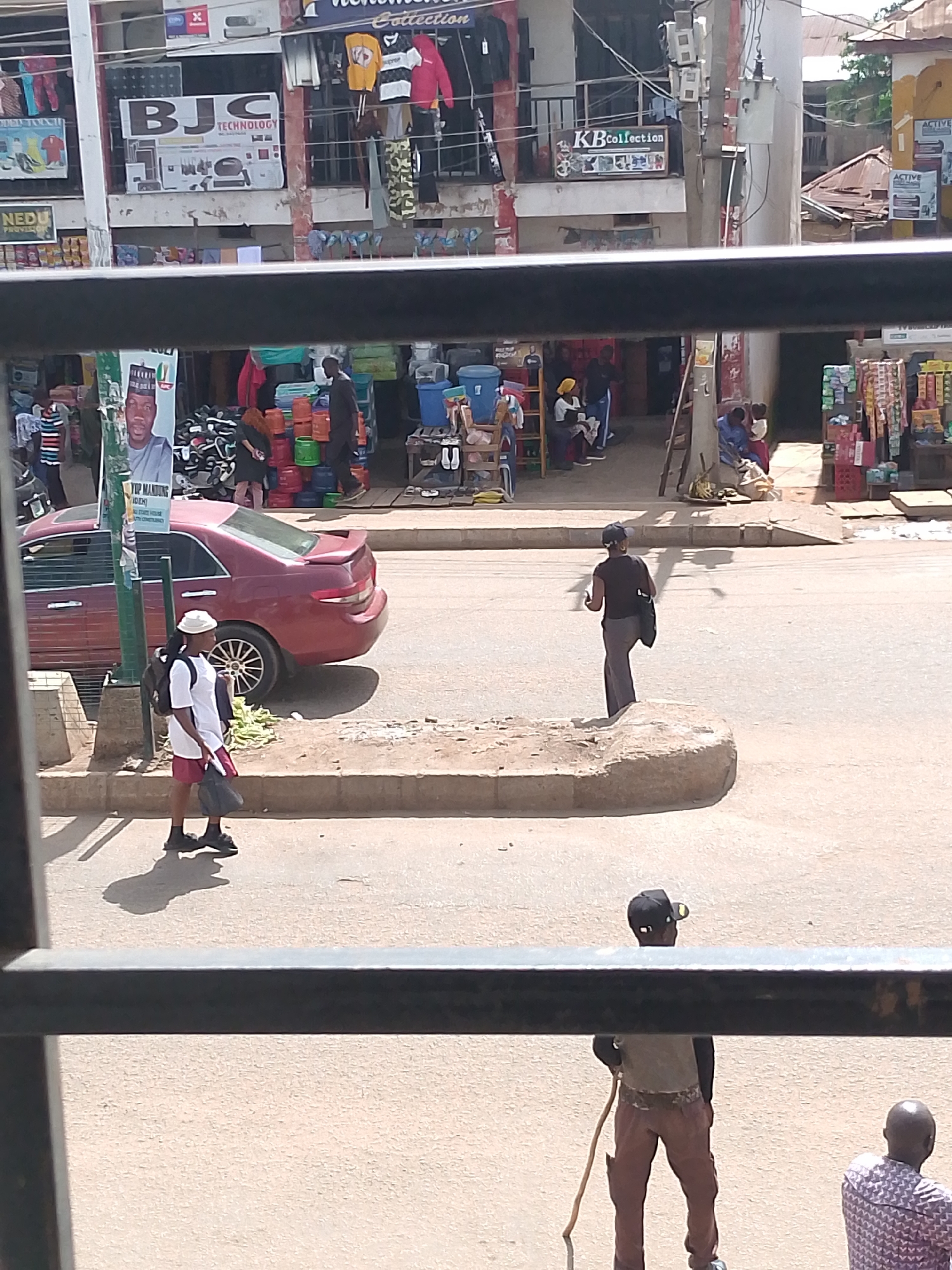 The image depicts a street scene in Bukuru, Nigeria. In the foreground, a person wearing a white t-shirt, red shorts, and a white hat is walking on the sidewalk, carrying a black bag. Behind them, a maroon sedan is parked on the road. Further down the street, a person in a black shirt and grey pants is standing on a concrete structure. In the background, shops are visible with various items displayed, including clothing and what appear to be household goods. Signs with text are present on the buildings, such as "BJC TECHNOLOGY" and "KBCollection." Other people are seen in and around the shops, suggesting a bustling market or commercial area. The lighting suggests it is daytime, and the weather appears to be clear.