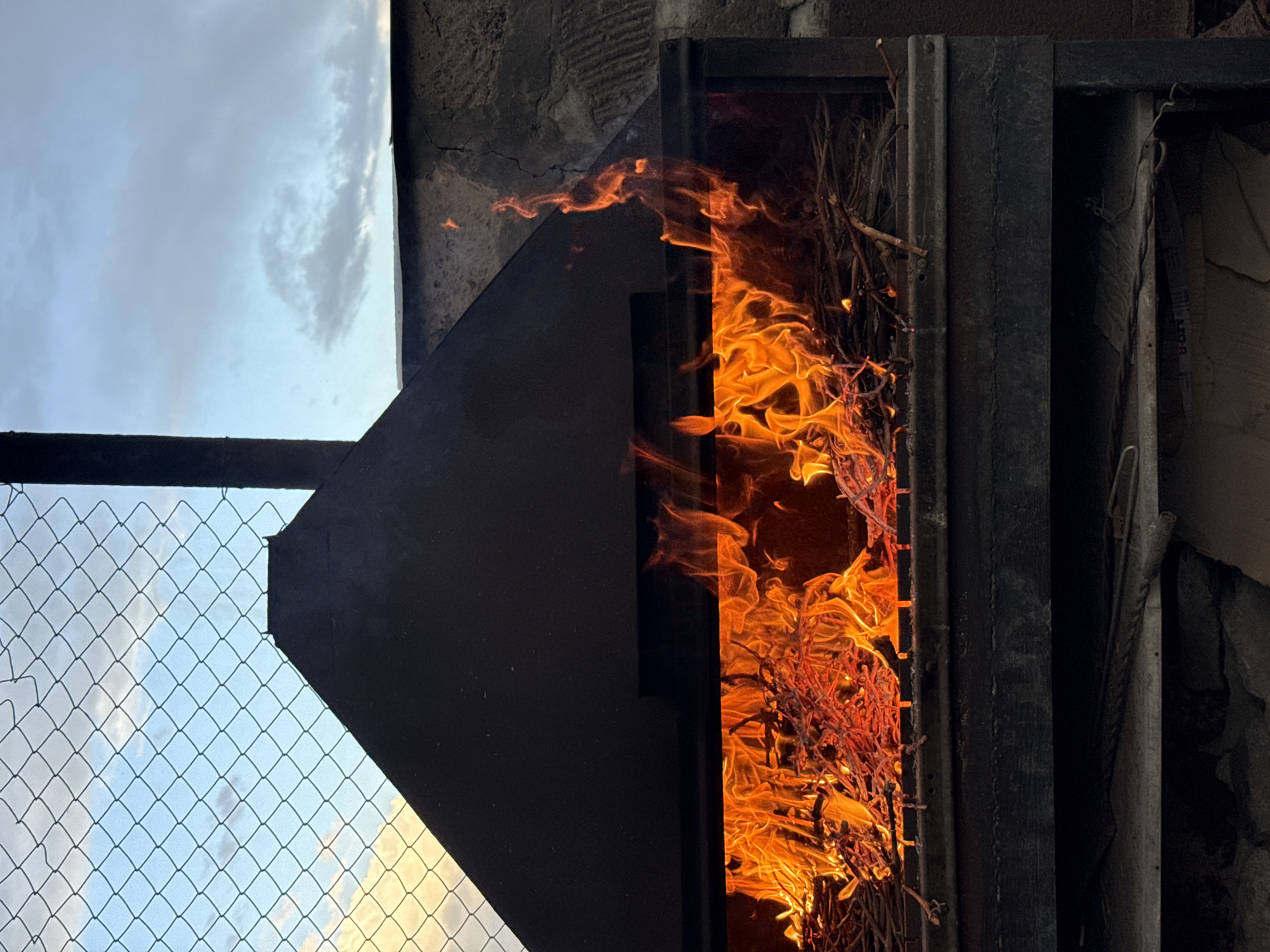 A metal structure containing a fire is the central element of the image, possibly located in Yerevan, Armenia. The fire is engulfing what appears to be twigs or small branches. To the left, a partially visible fence and a segment of sky are visible, hinting at an outdoor setting. The overall scene is framed by a stark contrast between the bright, intense flames and the darker, metallic structure, alongside the softer background elements.