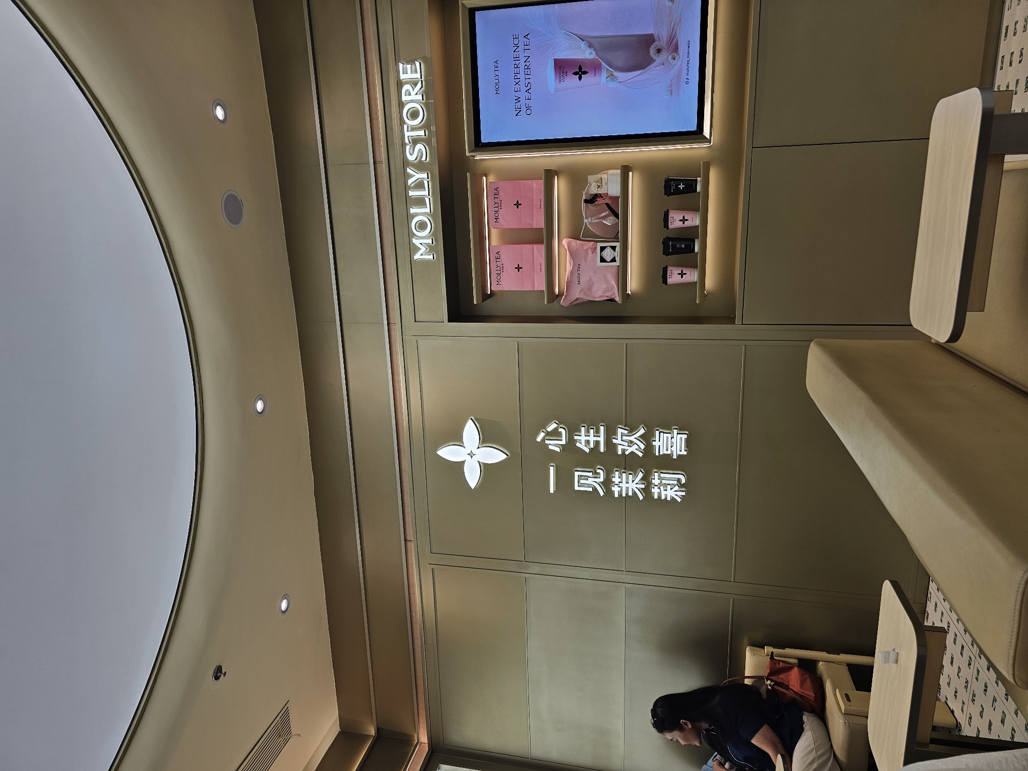 The image shows the interior of a "MOLLY STORE" in Condongcatur, Indonesia. The store features a beige and gold color scheme with a curved ceiling and recessed lighting. On the left side of the image, a woman with dark hair is seated on a beige chair, facing left. She is wearing a dark blue top and beige pants, and has a red handbag on the chair beside her. A small, light-colored side table is positioned in front of her.
The right side of the image displays the store's merchandise. A prominent sign reads "MOLLY STORE" in illuminated white letters. Below the sign, shelves display pink shopping bags and other products, including cups with pink and black designs. To the right of the shelves is a digital screen showcasing an advertisement for "MOLLY TEA" with the slogan "NEW EXPERIENCE OF EASTERN TEA." The advertisement features a stylized image of a tea cup and floral elements.
The floor has a patterned tile design. The overall environment appears to be a modern retail space designed for a tea shop.