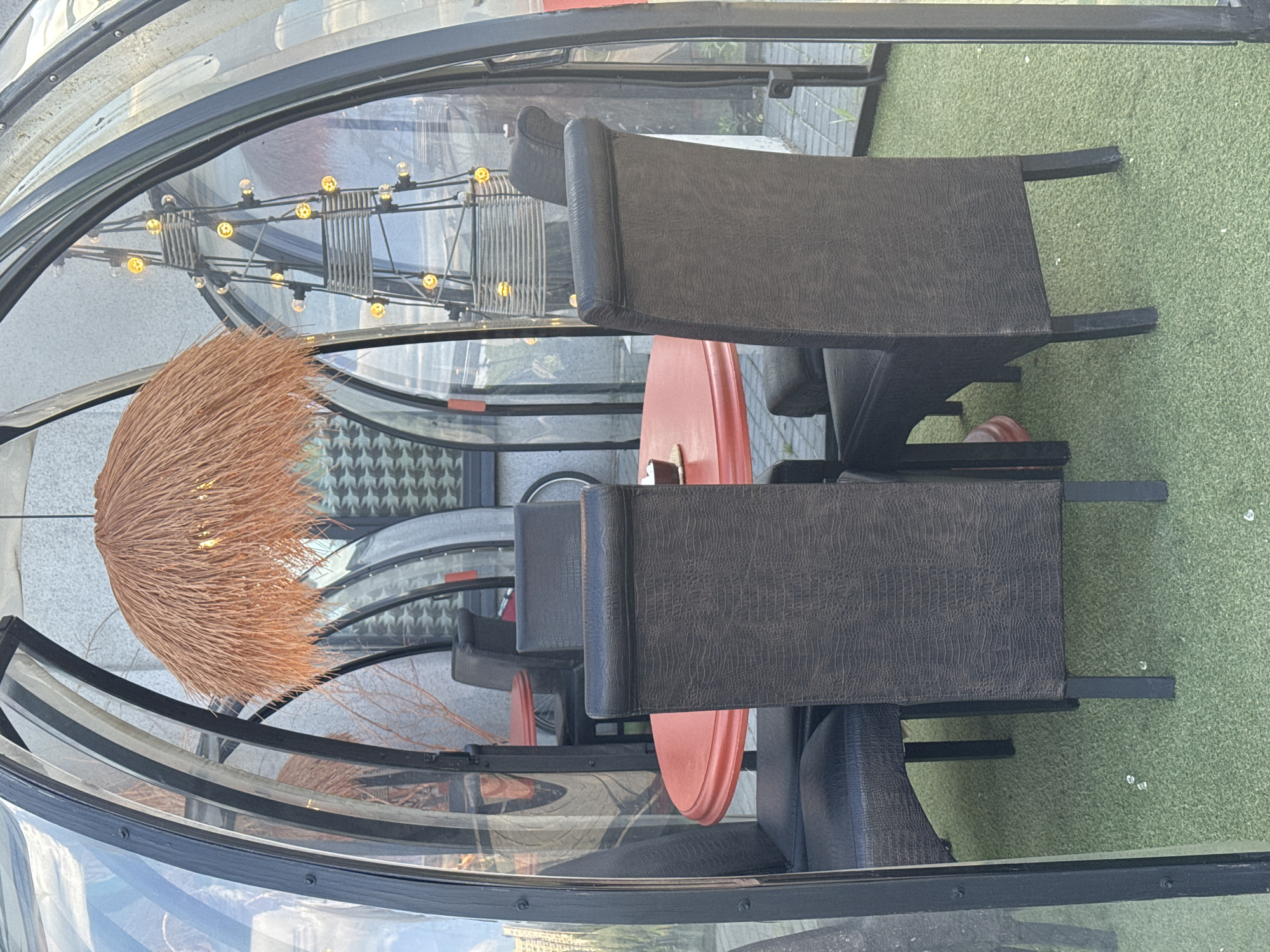 An outdoor dining area, part of a series of transparent, arch-framed enclosures, is depicted in Izhevsk, Russia. The specific enclosure shown features clear, curved panels supported by black metal structures, creating a 'pod' setting. Centrally positioned within the enclosure is a circular table with a reddish-orange top, upon which rests a small dark coaster and a light-colored object, possibly sugar packets or a napkin. Four high-backed chairs with dark upholstery, exhibiting a distinct textured pattern resembling crocodile or alligator skin, surround the table. The chairs have dark, straight legs.
Overhead, two distinct light fixtures are suspended: one large, dome-shaped fixture made of light brown, straw-like material, and another taller, sculptural fixture resembling a dark metal pyramid or ladder adorned with multiple spherical, yellow-glowing bulbs. The floor of the enclosure is covered with light green artificial turf, scattered with a few small white specks.
Through the clear panels, reflections of the surrounding environment are visible, along with parts of adjacent similar dining pods. In the background, a light gray, textured building wall is discernible, featuring a repeating dark geometric pattern on one section. Further elements in the exterior background include exposed bare branches of a tree or shrub, and portions of paved ground or sidewalk, with a bicycle wheel faintly visible on the far right. No people or active interactions are present in the scene.