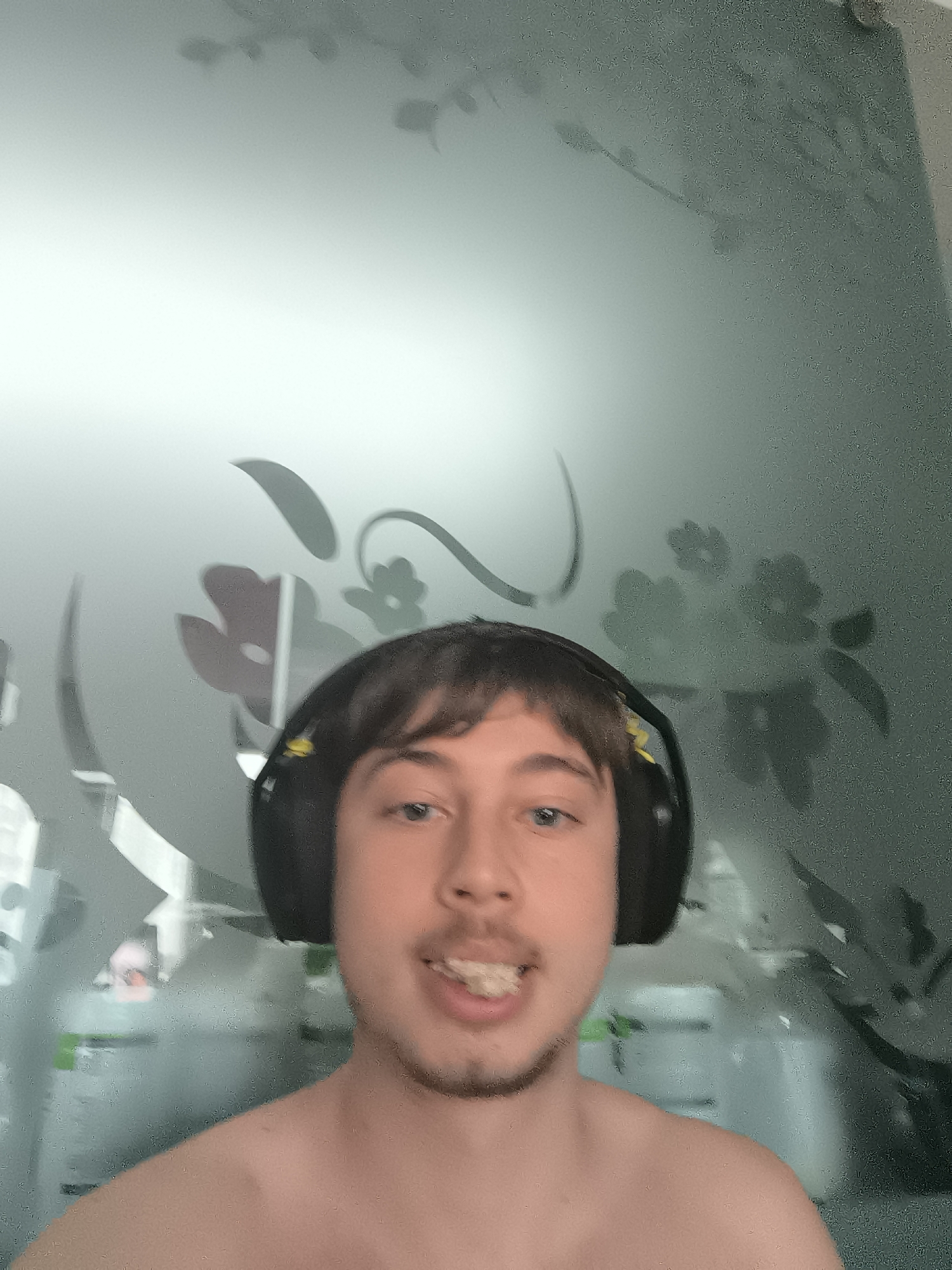 A young man is depicted in a close-up, likely a selfie or a video call screenshot, looking directly at the camera. He has short, dark hair, light-colored eyes, and a light goatee. He is shirtless and wearing black over-ear headphones, which have a small yellow detail visible on the left earcup. His mouth is open, revealing his upper teeth.
The background features a semi-transparent or frosted glass partition or wall with decorative etched patterns of leaves and flowers. Through the blurry glass, several large white containers are vaguely visible, suggesting an indoor setting such as a gym, a home workout space, or a room with storage. The lighting is bright and even, illuminating the man's face from the front. No discernible text is visible.