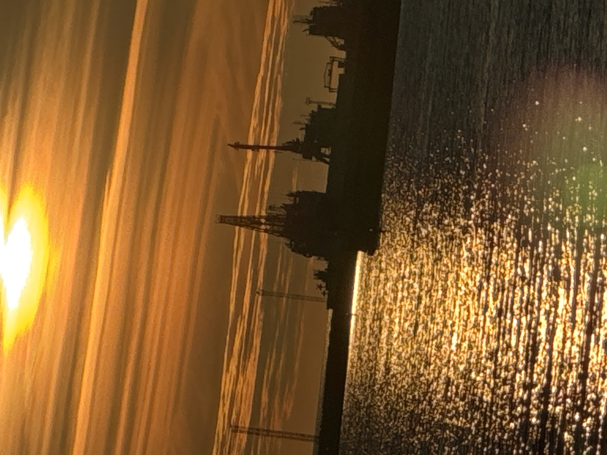 The image shows a sunset over the Tallinn Bay in Estonia. The sun is low in the sky, casting a warm, orange glow across the clouds and water. Silhouettes of ships and cranes are visible in the distance, suggesting an industrial or port area. The water is rippling, reflecting the sunlight in a shimmering pattern. The scene is peaceful and serene, with a sense of calm.