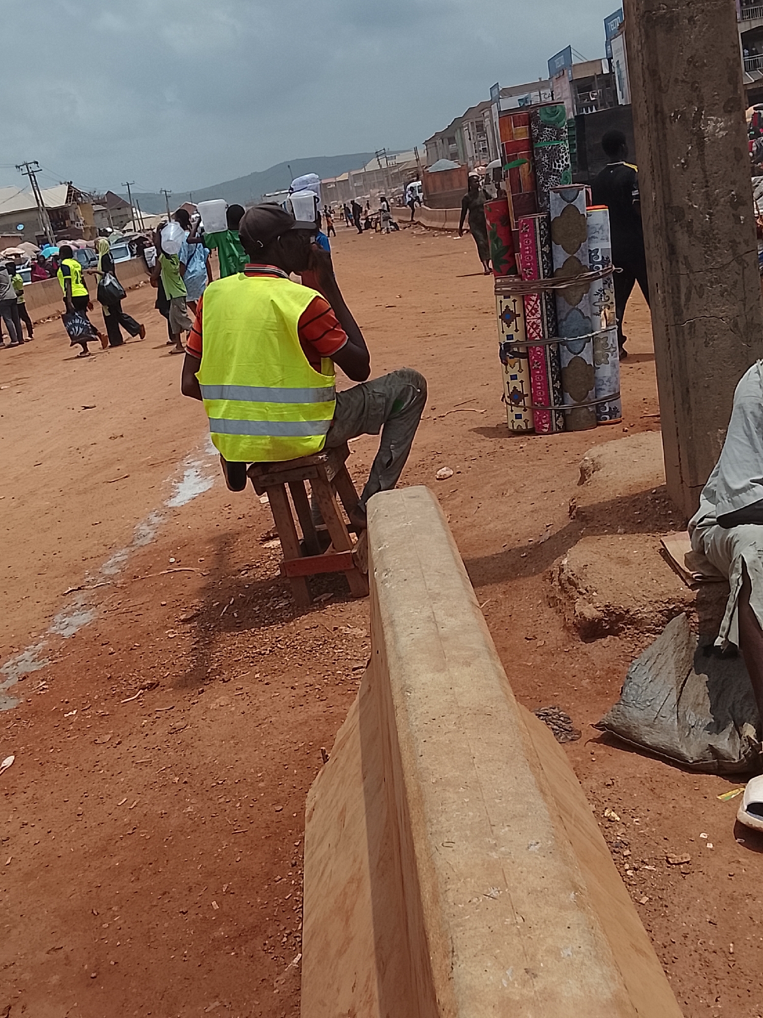 The image depicts a dusty street scene in Katambe, Nigeria. The ground is reddish-brown dirt. In the foreground, a man wearing a bright yellow safety vest and camouflage pants sits on a small, wooden stool, facing away from the camera, seemingly on the phone. A concrete barrier runs along the side of the street. In the background, several people walk in both directions, some carrying items on their heads. A stack of rolled-up rugs or mats tied together stands near a utility pole. Buildings are visible in the background, and the sky is overcast.