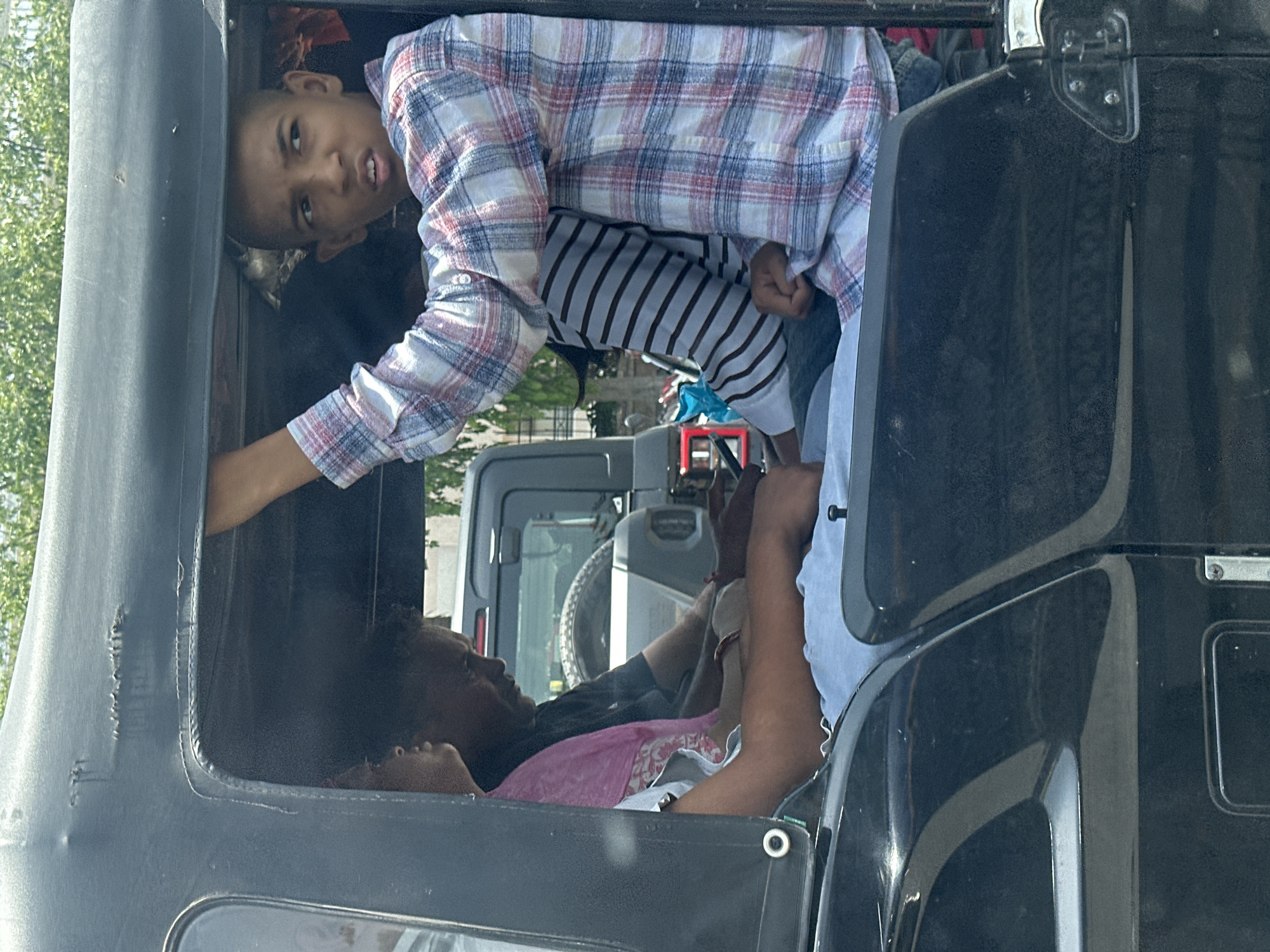 The image captures a scene inside a black auto-rickshaw in Ajmer, India. Three people are visible. In the foreground, a young boy with a shaved head is looking to the right, his mouth slightly open, and he is wearing a plaid shirt. To his left, an adult male with dark skin and short black hair is looking down, wearing a dark t-shirt. Between them, a person wearing a pink garment with a patterned hem is partially visible. The interior of the auto-rickshaw appears worn, with visible damage to the fabric roof. Outside the vehicle, a glimpse of trees and another vehicle, possibly a grey SUV, can be seen. The overall lighting suggests a bright, sunny day.