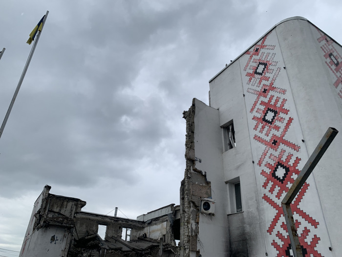 The image shows a damaged building in Derhachi, Ukraine, located at the coordinates 50.113, 36.107. The building's exterior is white with a large, red and black, geometric pattern covering the right side. There is a large section missing from the top of the building, and there are signs of damage throughout. Part of the roof is missing, exposing the interior of the building. There is a single, Ukrainian flag flying from a pole. The sky is overcast and gray. There are no people visible in the image. The scene appears to be a result of a bombing or other conflict-related incident.