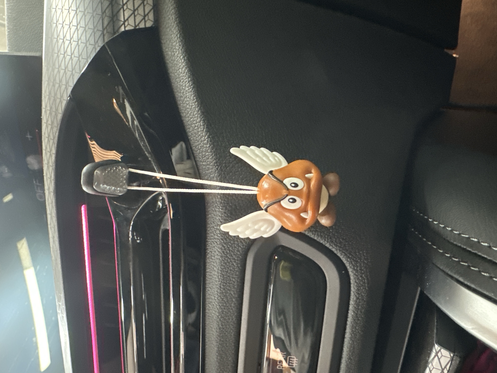 The image displays the interior of a modern car, with a focus on the dashboard and an air vent. A small, winged Goomba-like character charm, similar to those from the Super Mario franchise, hangs from a car air vent. The character is brown with large white eyes, an open mouth, and distinct white wings, suggesting it's a "Paragoomba." It is attached to a black car vent clip by a thin white string.
The car's dashboard features various textures, including a dark, pebble-grained material and reflective black plastic surfaces. To the left, a vertical pink-colored LED light strip illuminates part of the interior, casting reflections on the glossy trim. Further left, a portion of a digital display or control panel is visible, with faint white text including "OFF" and other indistinct icons. Below the hanging charm, a control button with the partially visible text "DEFR" (likely indicating a defroster function) is present. On the right side of the image, the dark leather upholstery of a car seat is visible, adorned with white contrast stitching. Above the vent, a textured panel with a geometric, grid-like pattern can be seen, possibly a speaker grille or decorative trim. The scene appears to be in low light, enhancing the visibility of the illuminated pink strip and reflections.
