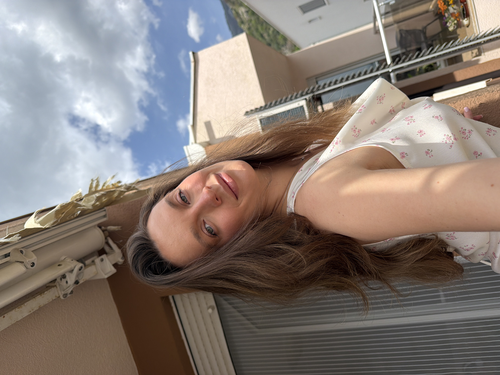 A young woman is featured in a close-up selfie, captured from a low angle. The original image is rotated 90 degrees clockwise. She has long brown hair, green eyes, and fair skin, with a subtle smile directed towards the camera. She wears a white top decorated with a delicate pink floral pattern.
When viewed correctly oriented, the background reveals a sunny daytime scene with a bright blue sky partially covered by scattered white and grey clouds. To her right, there are terracotta-colored building facades with balconies. One balcony in the upper right displays potted plants with vibrant orange and yellow flowers, while a window with horizontal blinds is visible below it. To her left, part of an outdoor awning or shutter mechanism, appearing weathered, is attached to a building structure. In the distance, lush green mountains suggest a scenic, possibly coastal, location, consistent with the provided context of Budva, Montenegro. The lighting indicates strong natural sunlight. There is no visible text in the image.
