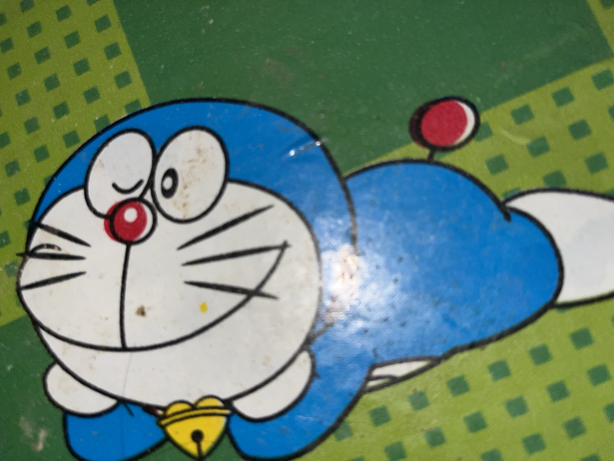 The image is a close-up of a cartoon character, Doraemon, lying on a green surface with a pattern of darker green squares. Doraemon is blue and white with a red nose and a yellow bell around its neck. The character appears to be smiling and winking. The background is a textured green surface. There are some smudges and marks on the surface, suggesting it may be worn or used. The image is well-lit, with no discernible shadows indicating a specific time of day. No text is visible in the image. The setting appears to be indoors, possibly a toy or a playmat. The location context provided is Benuaraja, Indonesia.
