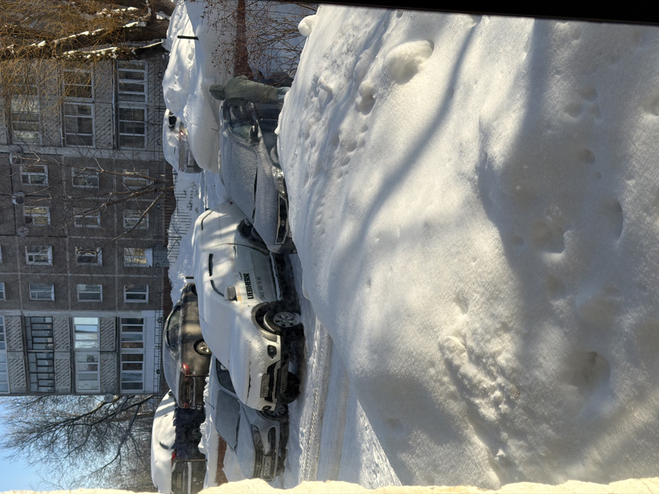 Cars parked on a snow-covered street line the side of a tall, Soviet-era building. The vehicles are coated in a thick layer of snow, obscuring their details. A person stands near the first car, likely clearing snow. A massive pile of snow looms in the foreground, dominating the scene and casting shadows. Bare trees and a clear blue sky are visible.