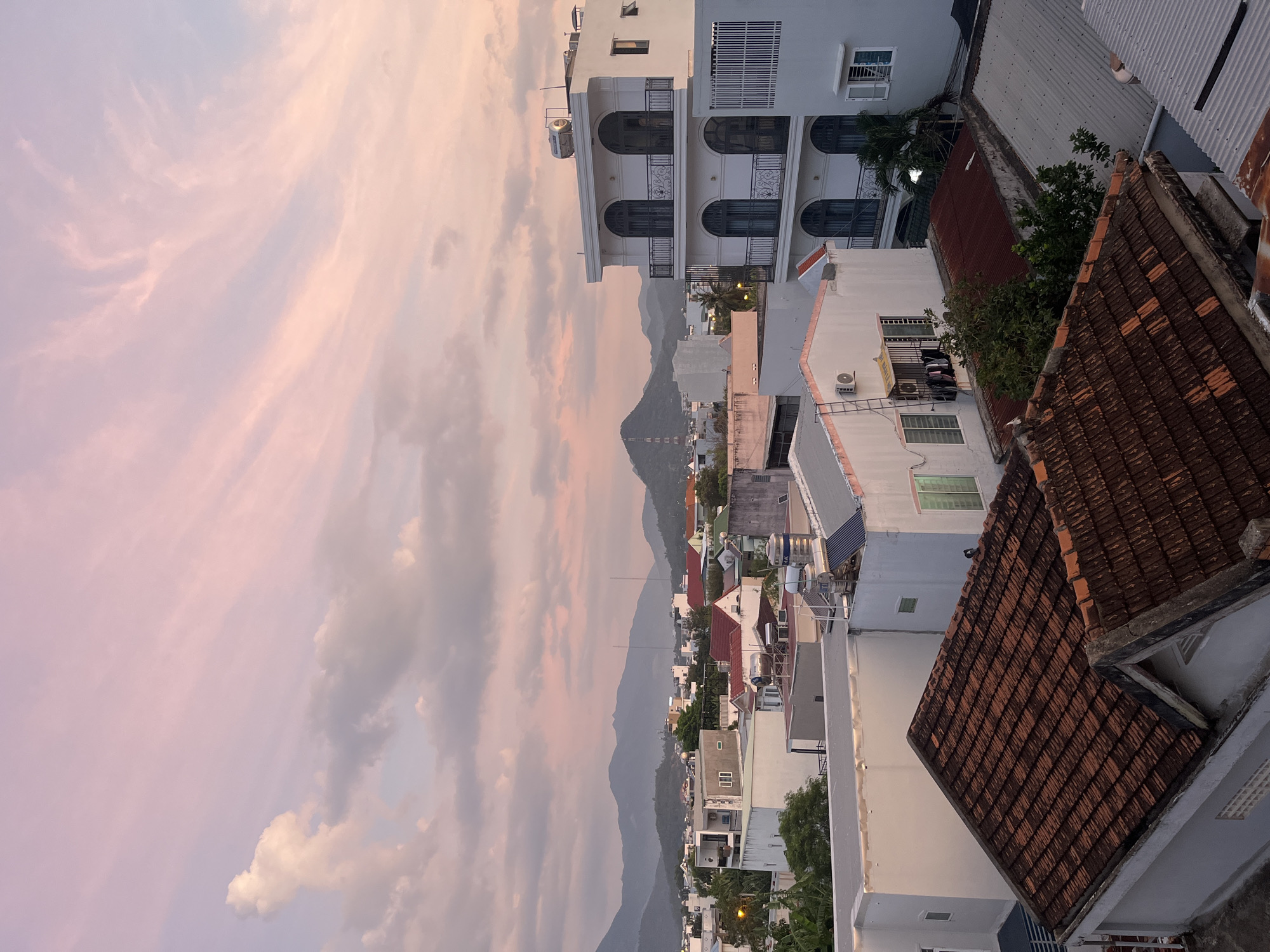 The image shows an elevated view of Bac Nha Trang Ward, Vietnam. The scene includes residential and commercial buildings of varying heights, predominantly white, with some displaying red tile roofs. A mountain range is visible in the background, obscured by a pink and grey cloudy sky, suggesting either sunrise or sunset. There are no people visible in the image. The environment suggests an urban setting bordering a natural mountainous landscape.