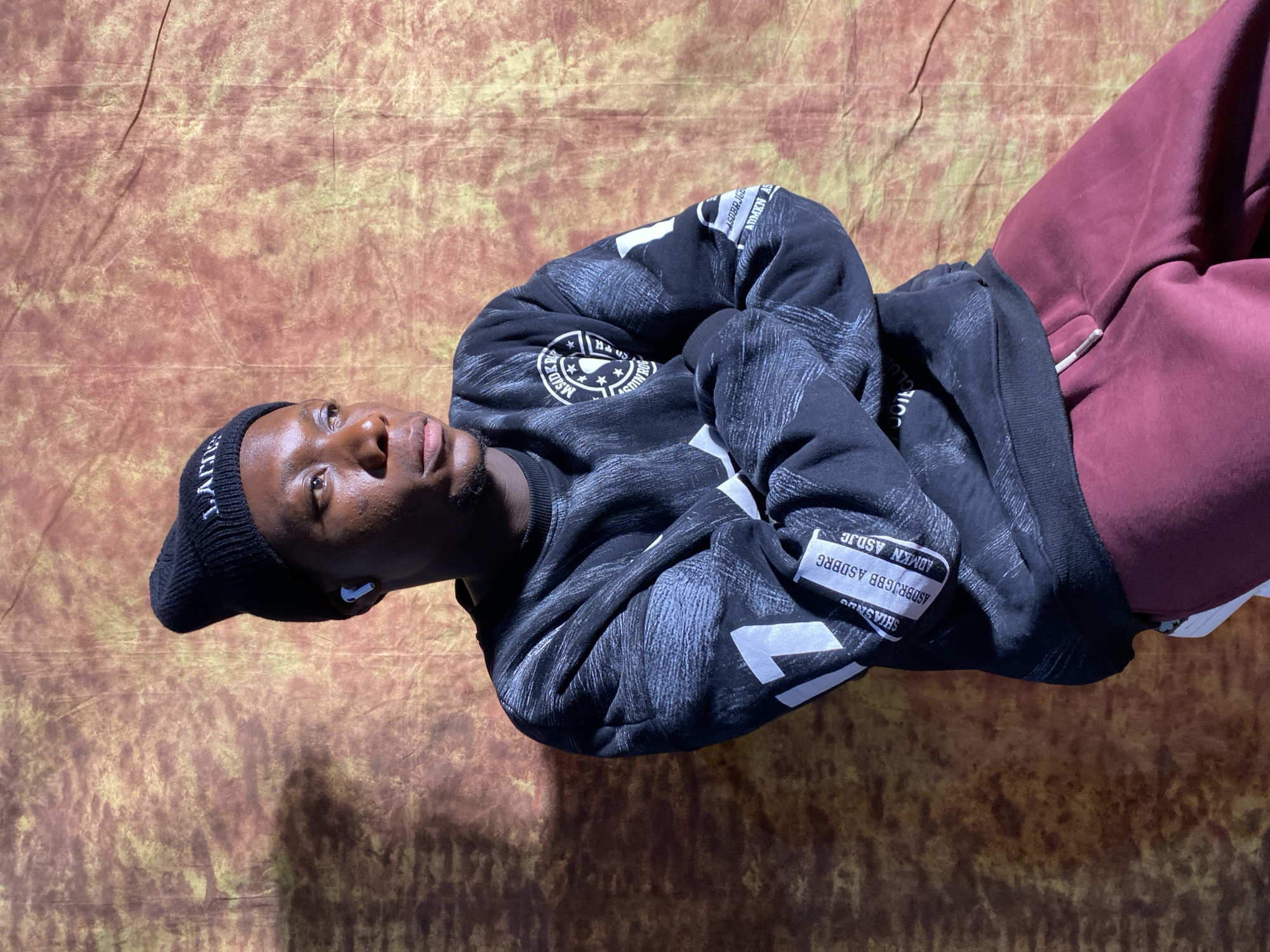 A young Black man is depicted in a portrait, seated and looking to his right with his arms crossed. He is wearing a black beanie, a black sweatshirt with a textured pattern and white graphics, and burgundy sweatpants. White earbuds are visible in his ears. The background is a textured fabric with warm hues of brown and yellow, casting shadows on the man. The image appears to be a studio portrait. There is no information in the image to suggest a location in Barakin Akawo, Nigeria.