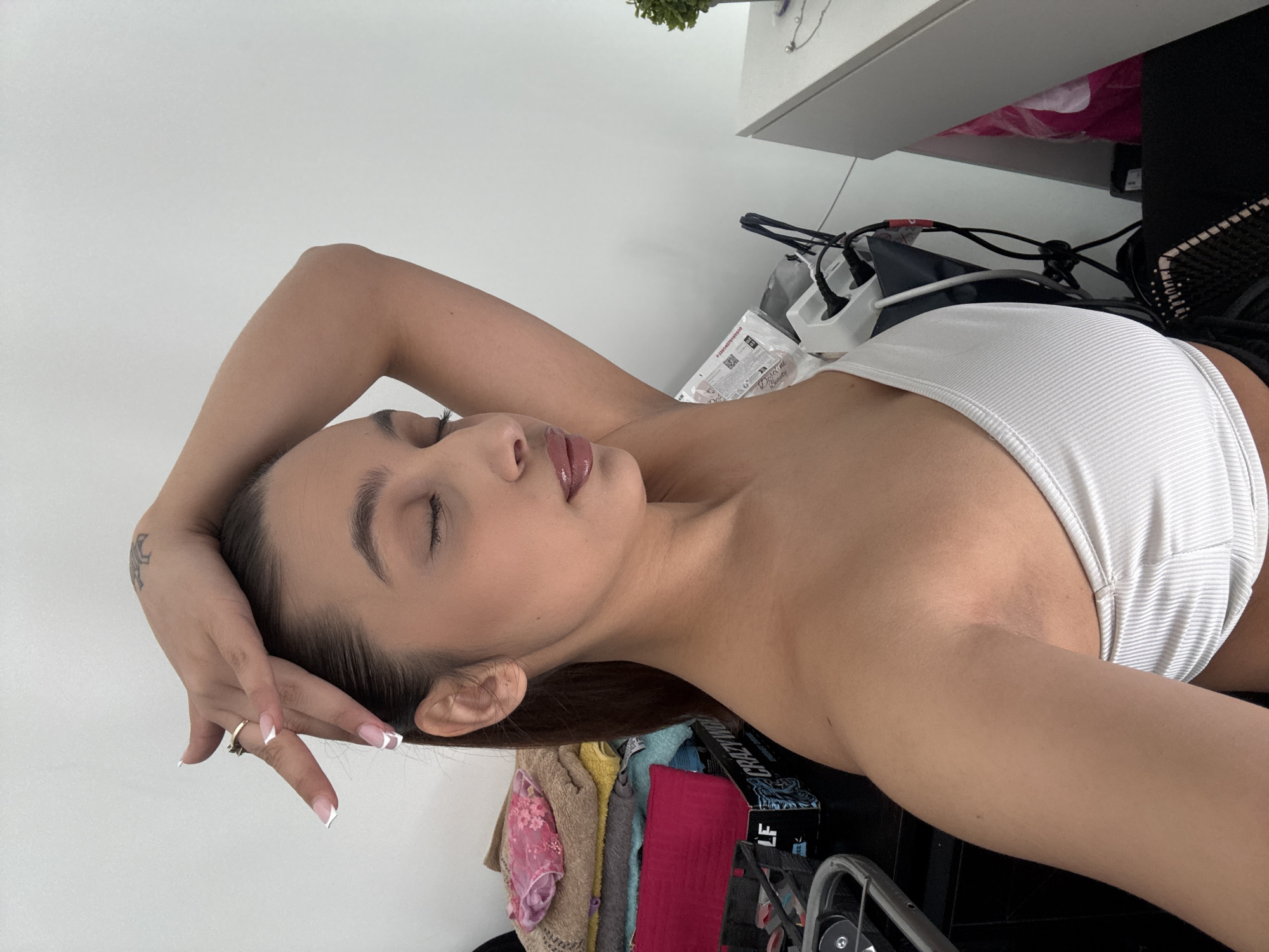 A young woman is depicted in a medium close-up, lying on her side with her eyes closed, suggesting a moment of rest or a posed photograph. She has dark hair, styled away from her face, and her lips appear glossy. She is wearing a white, ribbed strapless top. Her right hand is raised, supporting her head, showcasing manicured nails with white French tips and a gold ring on her ring finger. A small, dark tattoo, possibly a crescent moon or abstract design, is visible on her left forearm/wrist.
The background indicates an indoor setting, contextually located in Mönchengladbach, Germany. To the left and above her head, a plain white wall is visible. To her right and below, the scene is somewhat cluttered. A white shelf or desk holds a small green potted plant and a hanging silver necklace. Below this, various cables, wires, and power strips are visible, connected to unseen devices. A white multi-socket power extension is prominent. Piles of folded clothing in various colors (pink, yellow, red, blue) are stacked, possibly on a bed or a surface below the desk. A clear plastic package, partly obscuring a product, is visible near her left shoulder; text on it includes "KOSMETIKPRODUKTE" and "Beauty." A dark box with "CRAZY" and "LF" partially visible is also in the background. Bright, even lighting illuminates the scene, suggesting daytime.