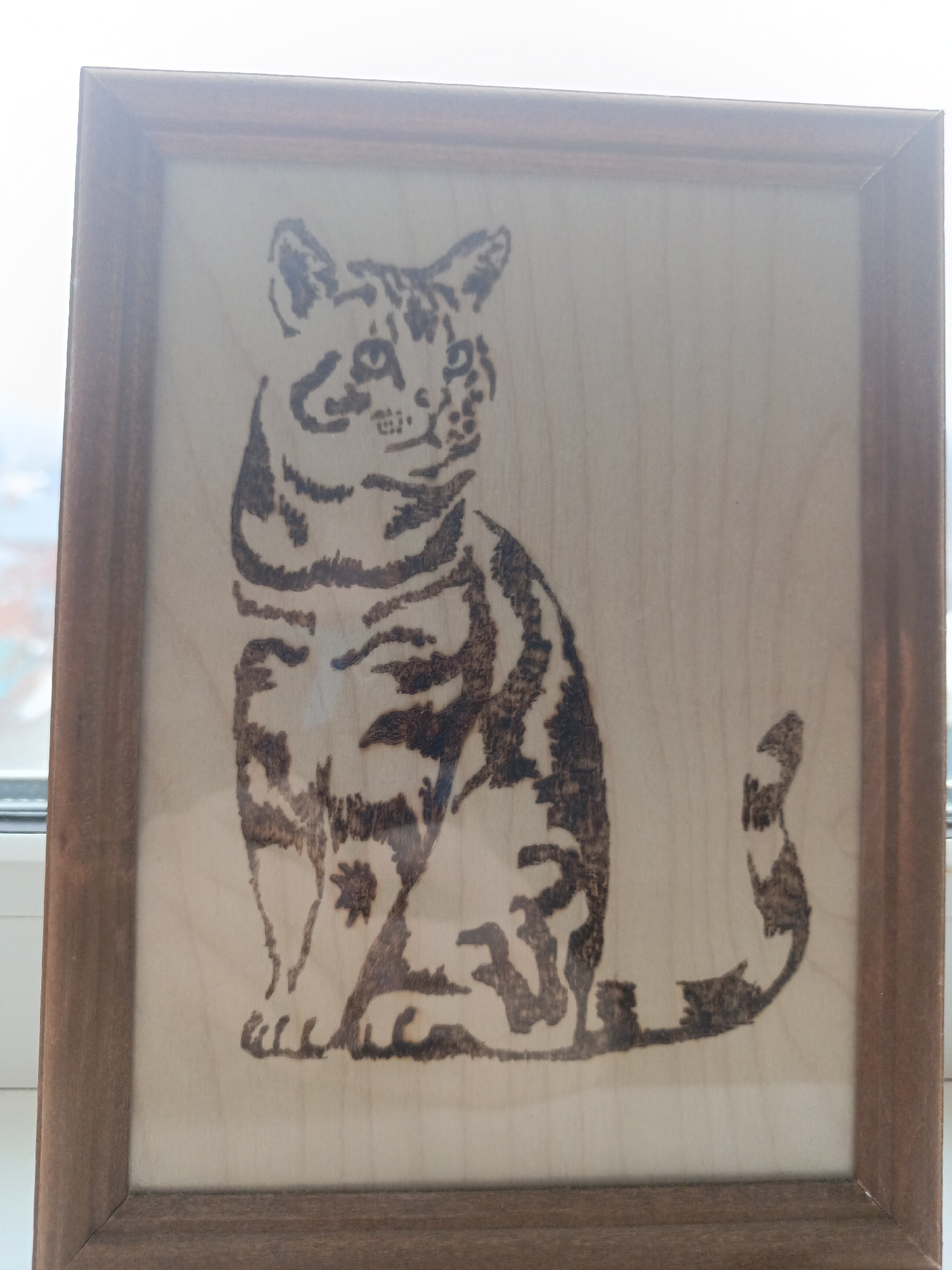 The image depicts a wood-burned artwork of a tabby cat in a wooden frame. The artwork is a light-colored piece of wood with a brown-burned image of a cat sitting upright. The cat has visible stripes and a curled tail. The frame is a simple, stained wood frame. The image appears to have been taken indoors near a window, with a blurred view of what might be buildings and sky visible through the window. The location is Chaikovsky, Russia. There are no other discernible people, actions, or interactions present in the image.