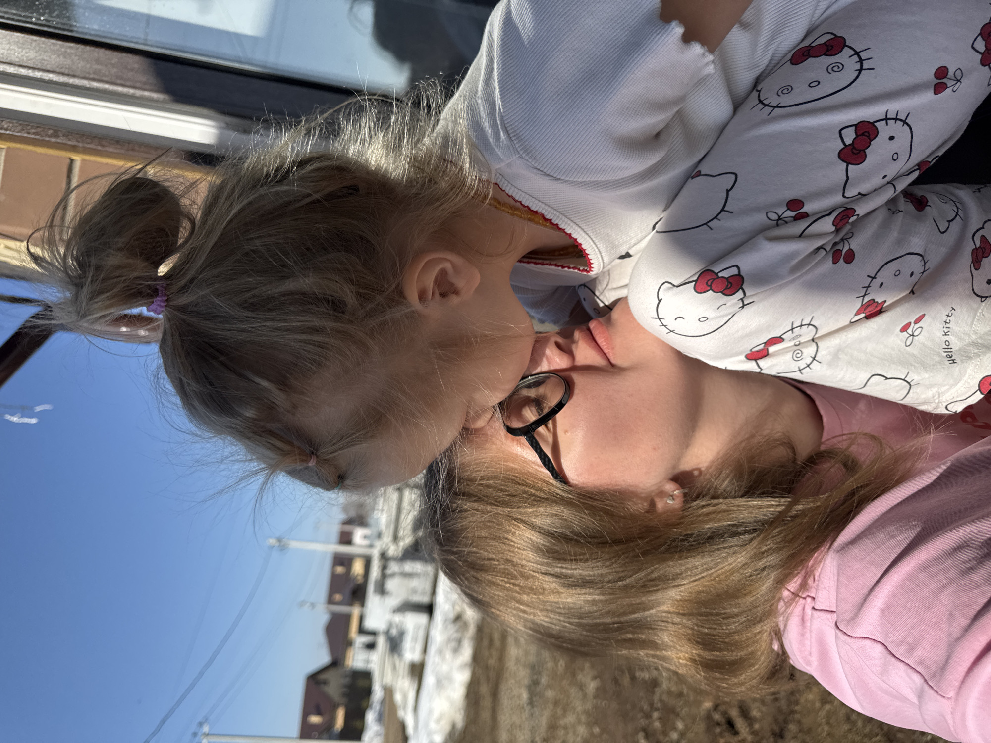 A young child is kissing an adult woman. The child has blonde hair pulled into a ponytail with a pink hair tie. They are wearing a white pajama top with a "Hello Kitty" pattern. The woman has blonde hair, wears glasses, and is wearing a pink shirt. The background shows residential buildings and snow on the ground, suggesting a location in a residential area of Gornyi, Russia, specifically Dvurechenskoe S/P.