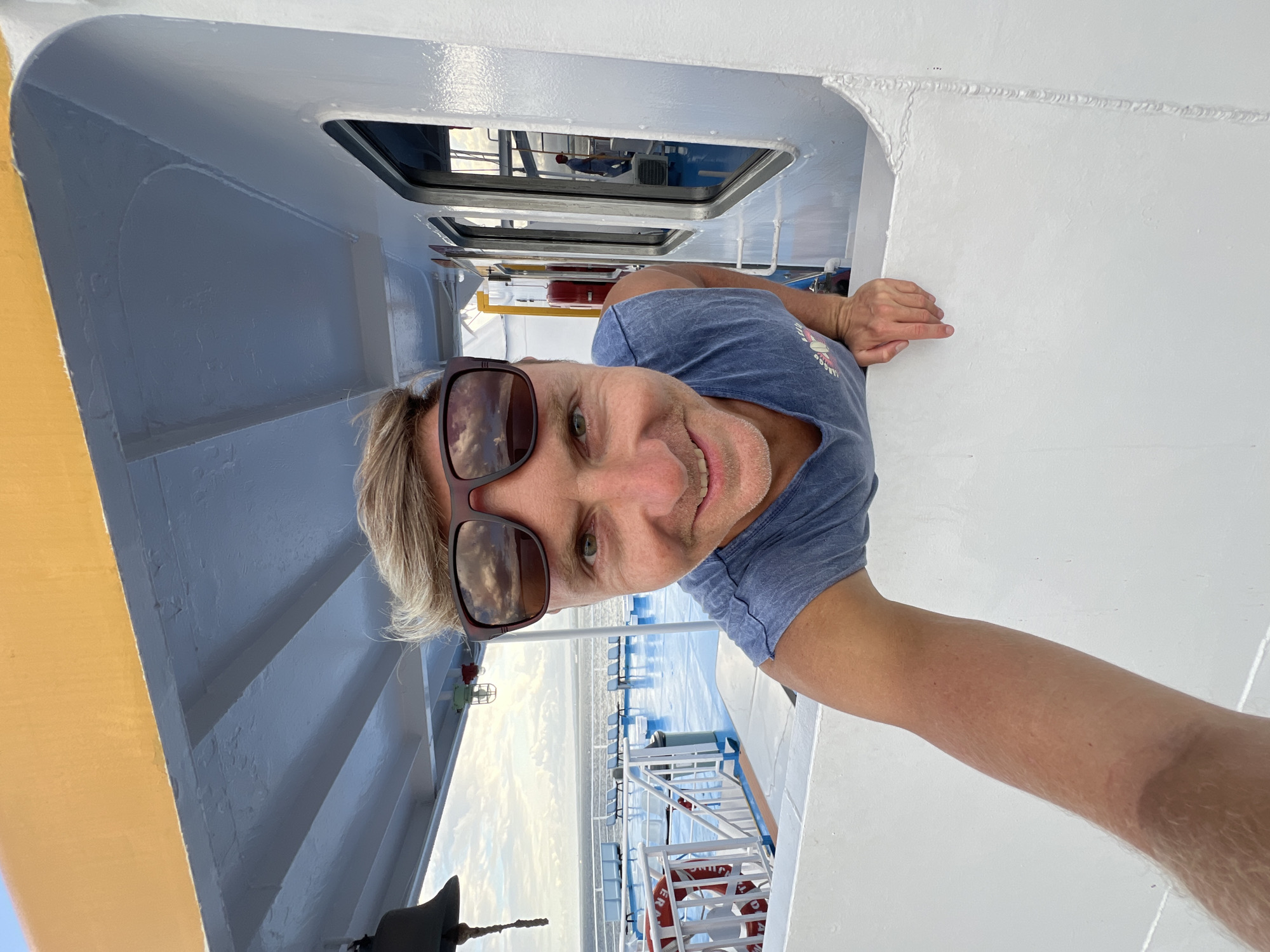 A man wearing sunglasses and a blue t-shirt is taking a selfie. He is on a boat or ferry, as indicated by the metal railings, life preserver, and view of the sea and coastline in the background. The man is looking at the camera with a smile. He is partially framed by a window or doorway on the boat. The location is Unknown, Indonesia.