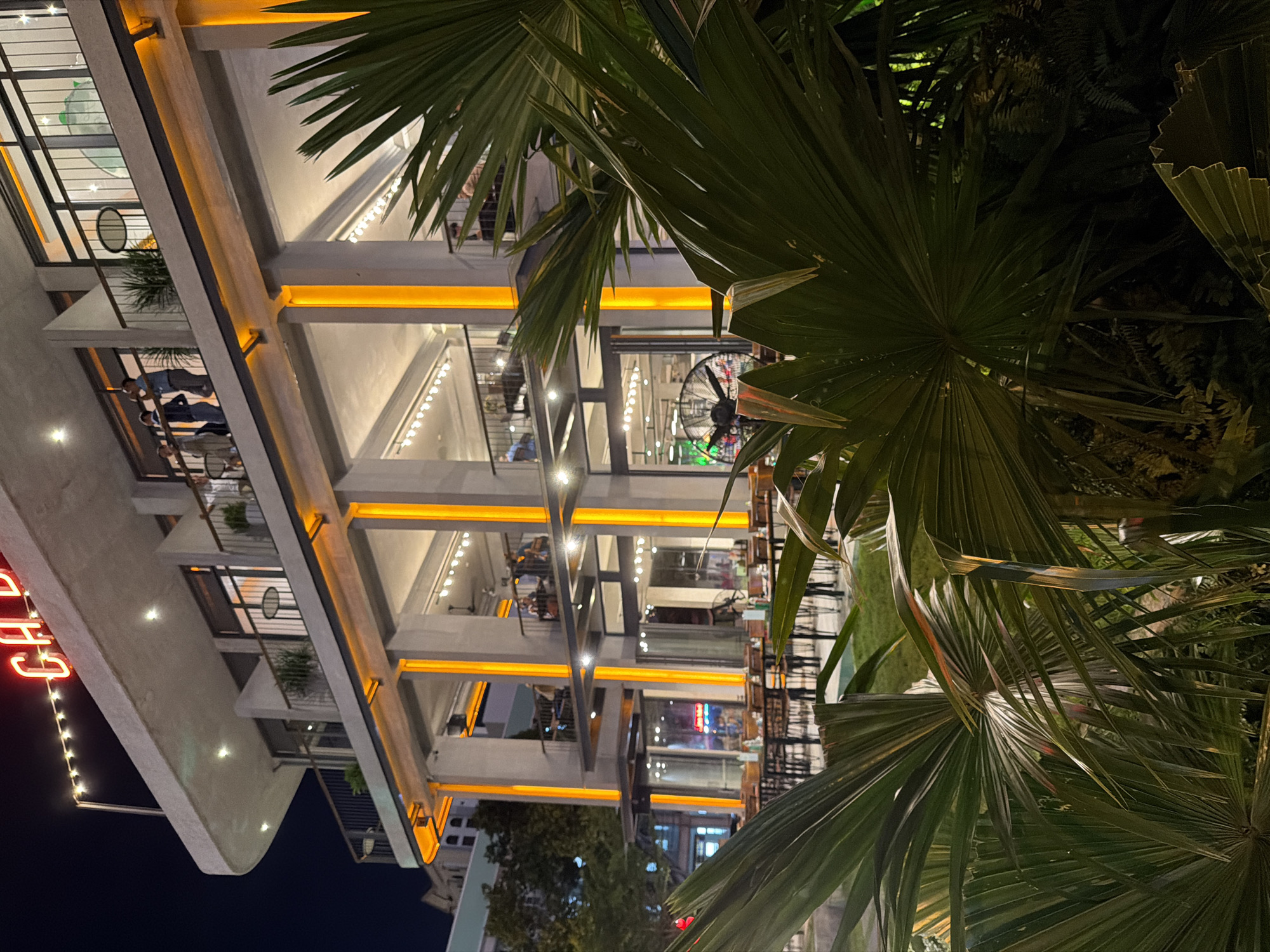 The image depicts a multi-story building, possibly a cafe or restaurant, at night in Nha Trang, Vietnam. The architecture features a modern design with concrete columns and floors, illuminated by warm orange strip lighting along the edges of the balconies and floors. Large glass windows allow a view into the interior spaces, where tables and chairs are visible, suggesting a dining or seating area. Small plants are placed on the balconies.
In the foreground, large, dark green palm fronds obscure parts of the building, creating a sense of depth and suggesting an outdoor seating area or a lush environment surrounding the establishment. The lighting is artificial, with yellow and white lights from within the building and on decorative strings outside. The sky is dark, indicating it is nighttime.
There are glimpses of people inside the building, appearing as silhouettes or indistinct figures, engaged in activities such as dining or socializing. A large industrial fan is visible through the glass on one of the upper floors. A neon sign with red lettering is visible on the left side of the building, partially obscured by the palm fronds. The text appears to read "Cafe." The overall atmosphere is one of a lively urban evening, with the illuminated building standing out against the night sky, framed by the tropical foliage.