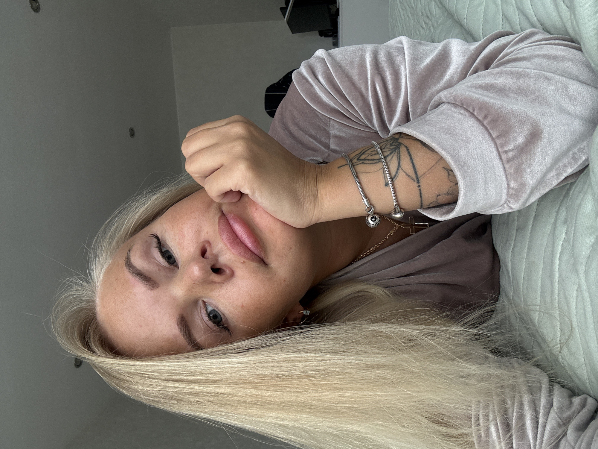 A woman with blonde hair and fair skin is lying down. She has freckles on her face. She is wearing a pink velvet top and jewelry on her wrist and neck. A tattoo is visible on her left forearm. The background includes a wall and bedding.