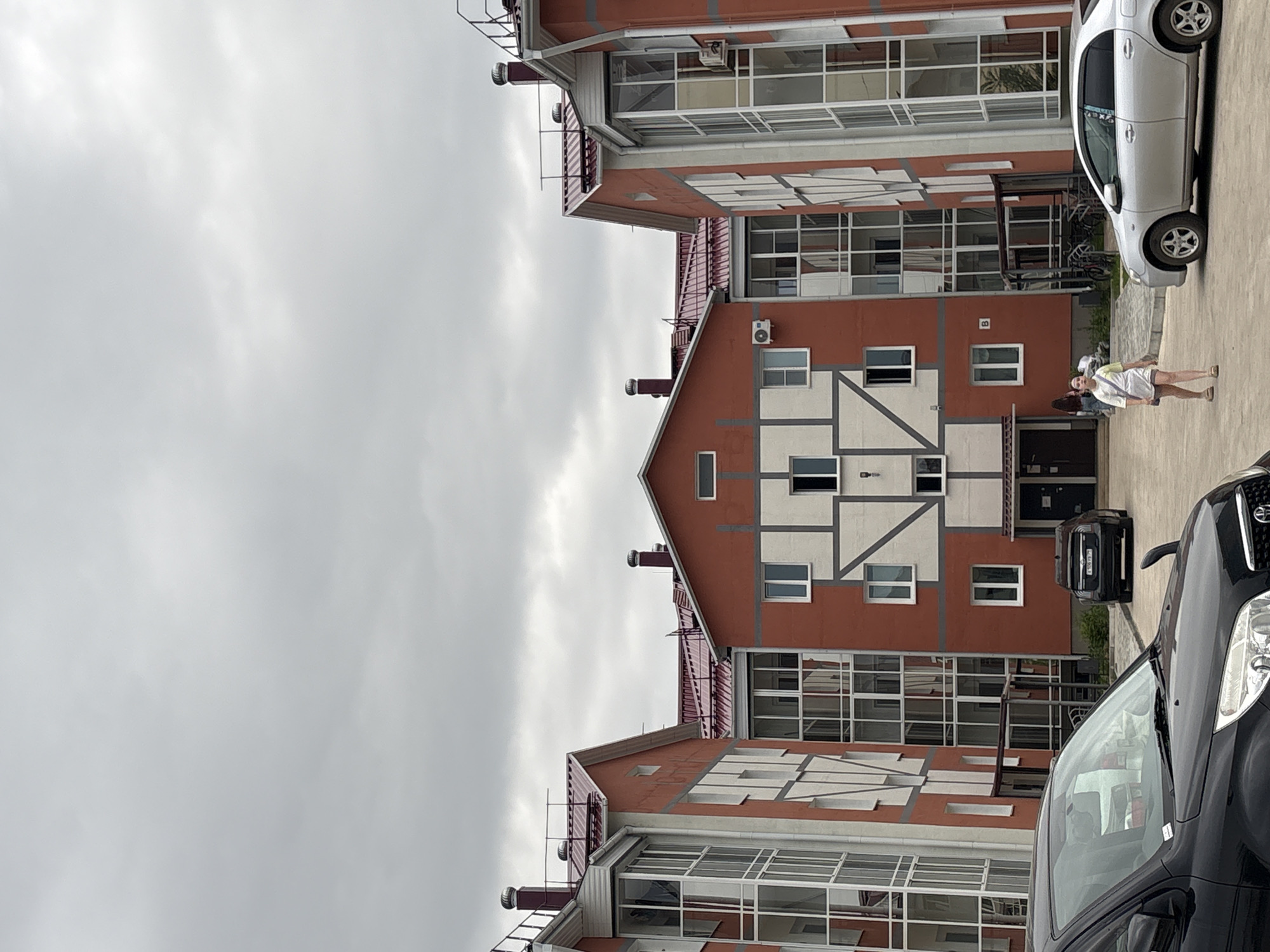The image shows a street scene in Birobidzhan, Russia. The focal point is a three-story residential building with a distinctive architectural design. The building is predominantly red-brown with white accents framing the windows and creating a decorative pattern on the central section of the facade. Each floor features numerous windows, with the top floors having larger, grid-like windows. A red roof caps the structure.
Two cars are parked on the pavement in front of the building: one is silver and another is black. Near the entrance of the central building, two individuals are visible. One appears to be facing away from the camera, and another appears to be walking with an outstretched arm.
The sky is overcast.