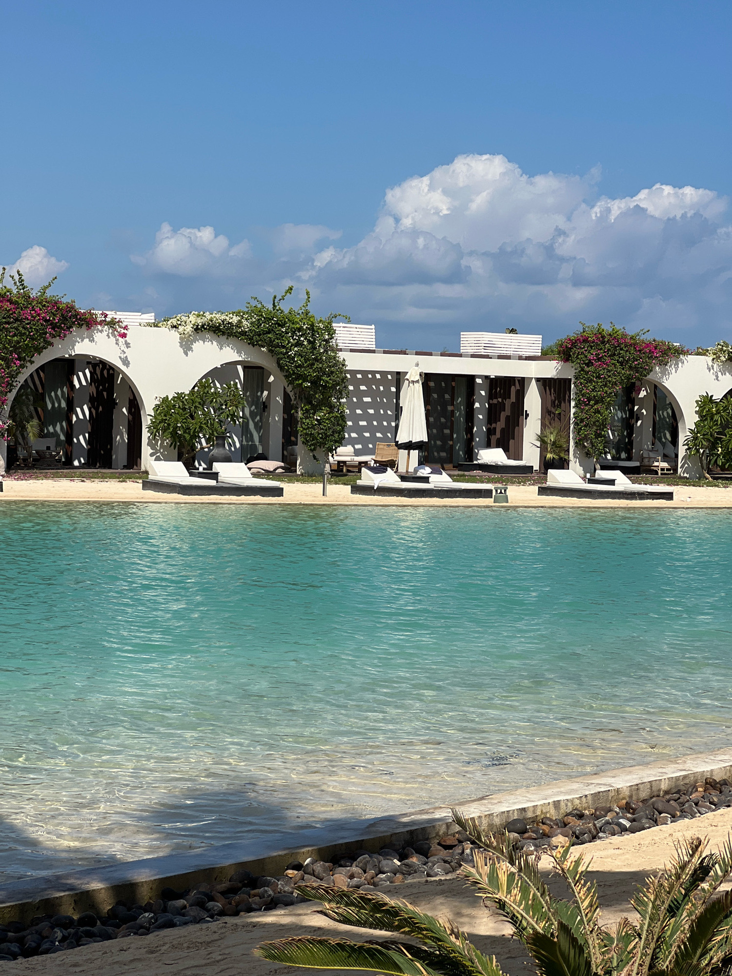 The image depicts a luxurious resort complex situated in Sidi Abdel Rahman, Egypt. The scene is dominated by a crystal-clear turquoise swimming pool in the foreground, with its shallow end revealing the sandy bottom and small pebbles lining the edge. Beyond the pool, a pristine sandy beach separates it from a row of white, arched structures that appear to be guest rooms or villas. These structures are adorned with vibrant pink and white bougainvillea cascading over their white walls. Multiple white lounge chairs are strategically placed on the sand in front of each arched entrance, inviting relaxation. Some of the entrances are partially obscured by decorative white lattice work and tall potted plants. The sky above is a brilliant blue, dotted with fluffy white clouds. The overall impression is one of serene opulence and tropical beauty.