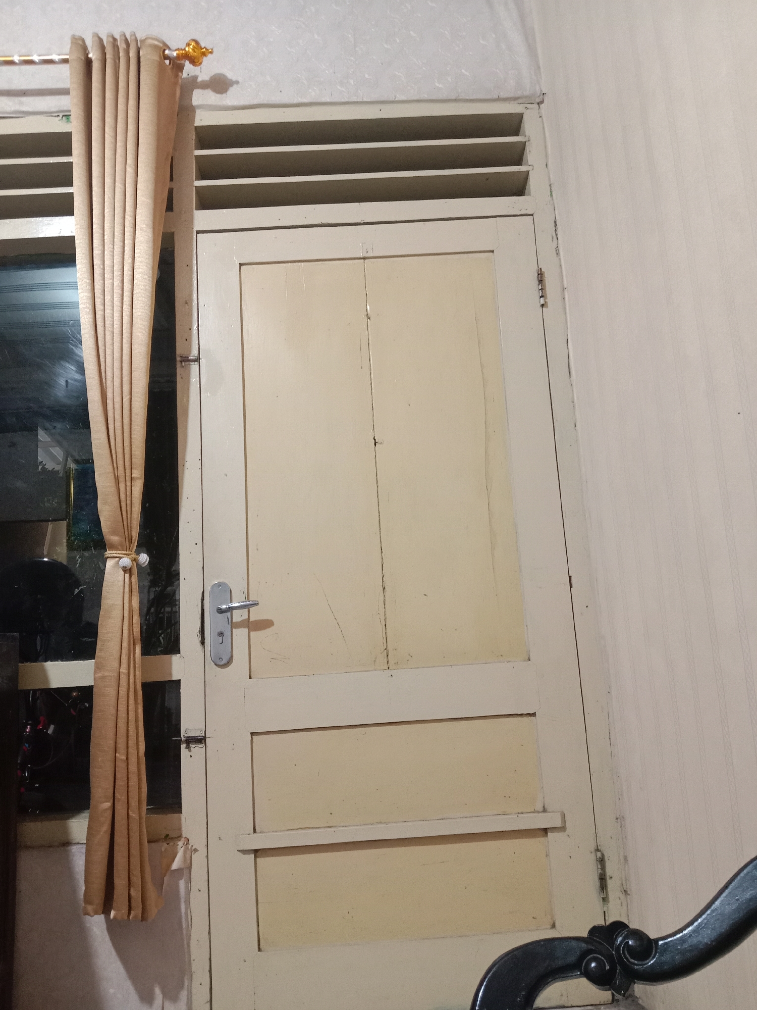 The image depicts an interior scene, likely a doorway, in Madiun, Indonesia. A closed, light-colored wooden door occupies the center of the frame. To the left of the door, a window is partially obscured by a golden-brown curtain, which is held back by a decorative tie. The curtain rod and finials at the top of the window suggest a domestic setting. Above the door and window, a louvered vent is visible. The wall to the right of the door has a textured, striped wallpaper. A metal door handle and lock mechanism are prominent on the door. In the lower right corner, a portion of a dark-colored, ornate metal chair or furniture piece is visible. The lighting in the scene appears to be artificial and somewhat dim, casting shadows.