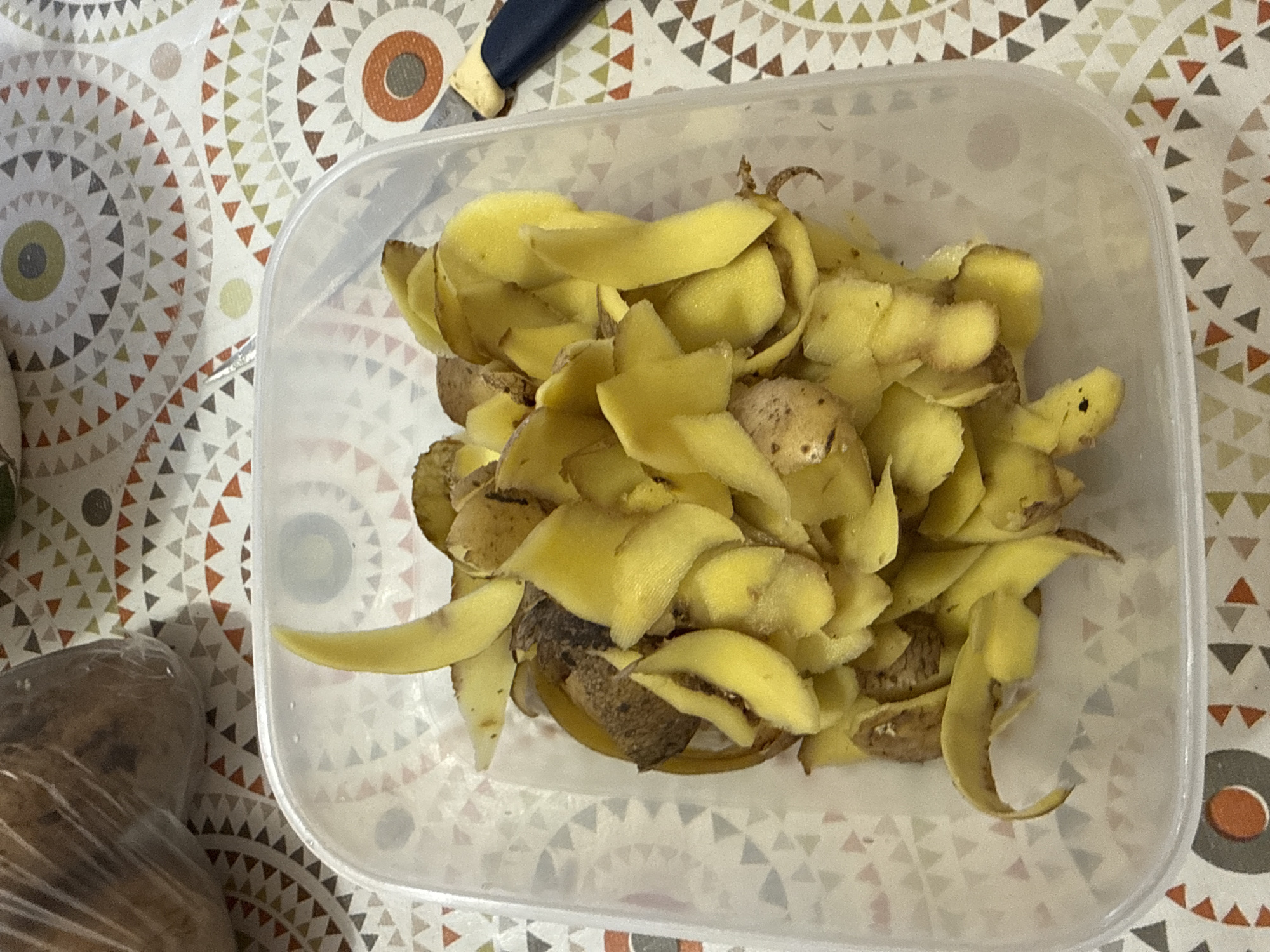 A clear plastic food container filled with potato peels is shown in the center of the image. The peels are yellow with some brown spots. The container is on a tablecloth with a geometric pattern of triangles in shades of orange, brown, and green. To the right of the container, the handle of a blue and yellow knife is visible. In the upper left corner, a blurry image of potatoes in a plastic bag can be seen. The image is taken from a top-down perspective. There is no visible indication of the city of Морозовка, Russia in the image.