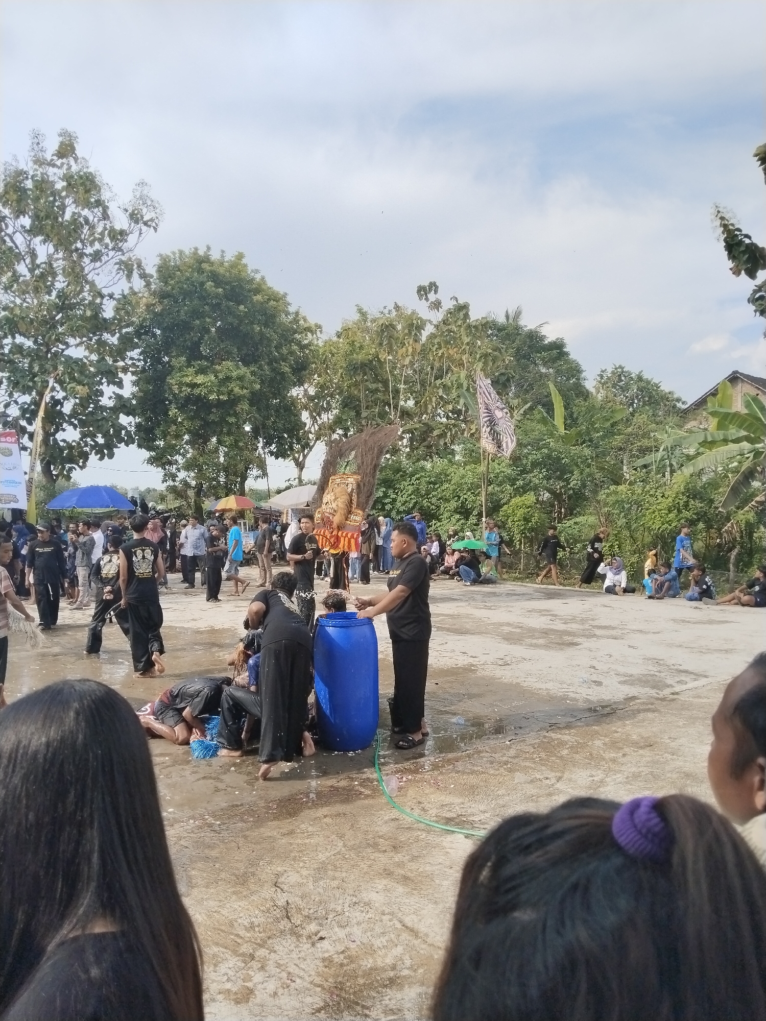 The image shows a scene in Mojodoyong, Indonesia. A group of people are gathered on a concrete surface. Several individuals in black clothing are attending to two people lying on the ground. A blue barrel is present, and a hose appears to be connected to it, with water being dispensed onto the people on the ground. The crowd includes people of different ages, some standing and others sitting. In the background, there are trees, tents, and buildings. A flag with a black and white pattern is also visible. The sky is partly cloudy.
