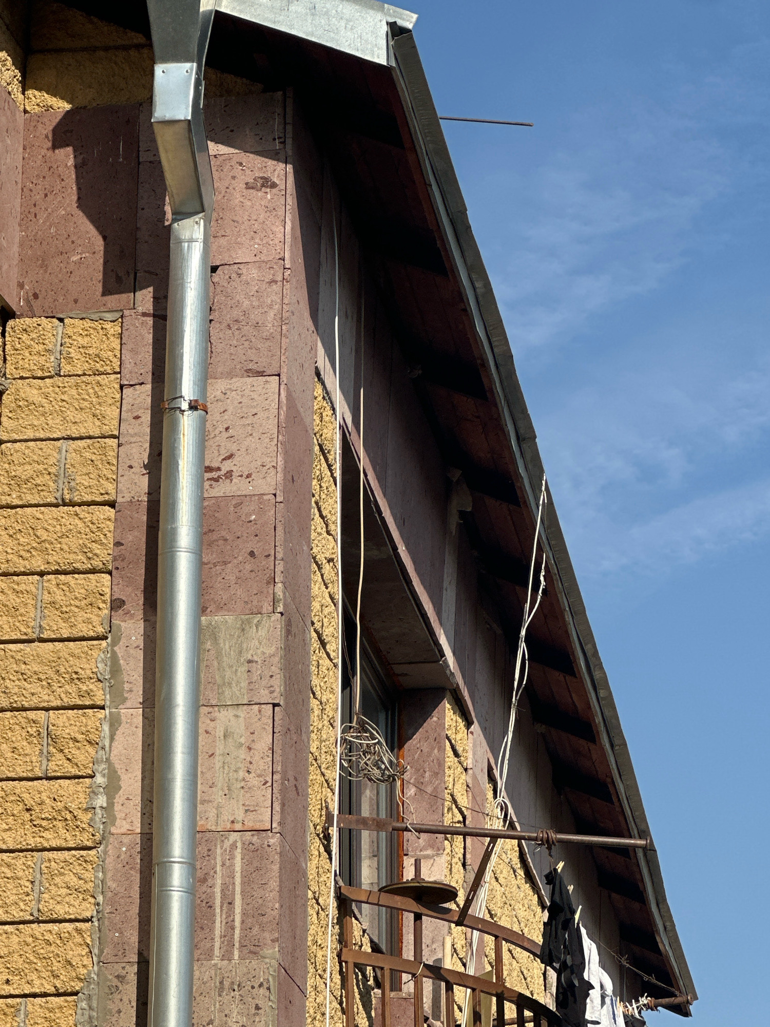 The image captures a side view of a building in Gyumri, Armenia, under a clear blue sky with wispy clouds. The building features a facade with two distinct textures: a section of rough-hewn yellow stone blocks and another section of larger, dark reddish-brown stone blocks, possibly basalt. A silver-colored downspout runs vertically along the corner where these two materials meet, connected by a horizontal bracket. The roofline is visible, with dark wooden eaves and a dark-colored roof. A rusty metal balcony railing is present, from which several articles of clothing are hanging on a clothesline, including a black t-shirt and a white garment. A circular metal object, possibly a small dish or a part of a ventilation system, is attached to the balcony structure. A tangled mass of white wire or cable is visible near a window above the balcony. A thin, horizontal metal rod protrudes from the building near the roofline. The overall scene suggests a residential building on a sunny day.