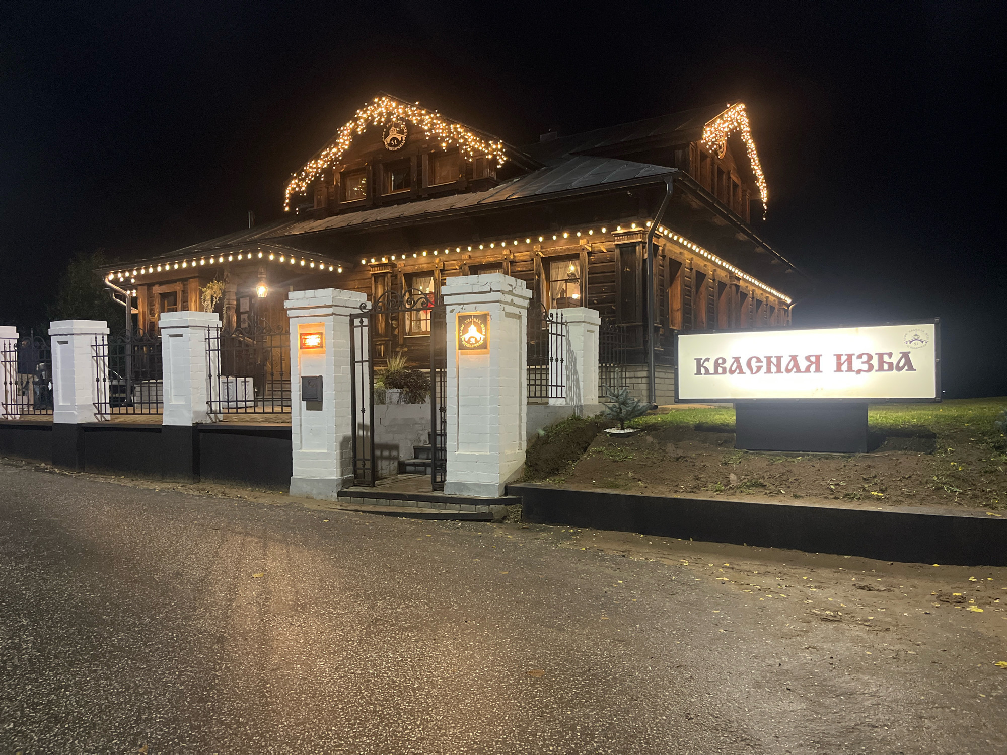 The image depicts a large, wooden, two-story building at night, likely a restaurant or guesthouse, in Suzdal, Russia. The building is illuminated by numerous warm-toned string lights adorning its rooflines and eaves, creating a festive atmosphere. A large, illuminated sign in front of the building displays Cyrillic text that reads "КРАСНАЯ ИЗБА," which translates to "Red Hut" or "Red Log House." The building has a traditional Russian architectural style with visible wooden logs and decorative window frames. A black metal fence with white decorative pillars encloses the entrance area. The ground in front of the building is paved with asphalt, which appears wet, suggesting recent rain. The surrounding environment is dark, with a hint of green grass visible behind the sign and to the right of the building. No people are visible in the image.