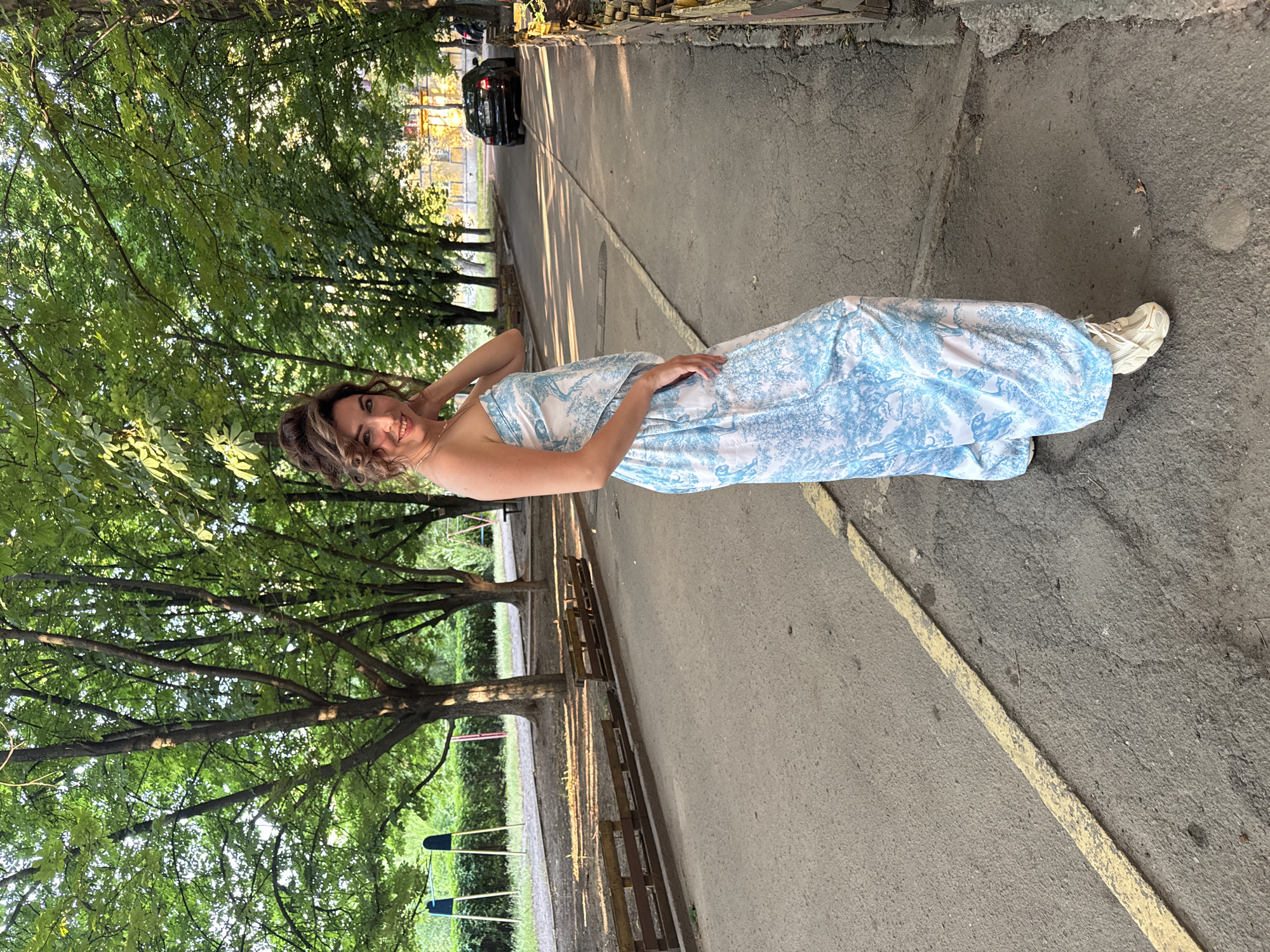 A woman in a blue and white patterned strapless maxi dress and beige sneakers stands on an asphalt path lined with trees. The woman is facing the camera, smiling, with her right hand on her neck. Behind her, a line of wooden benches runs along the path, and further back, there is a playground with swings. A black car is parked on the street in the background, with a building visible behind it. The scene appears to be a public park or residential area. The location is the city of Kryvyi Rih, Ukraine.